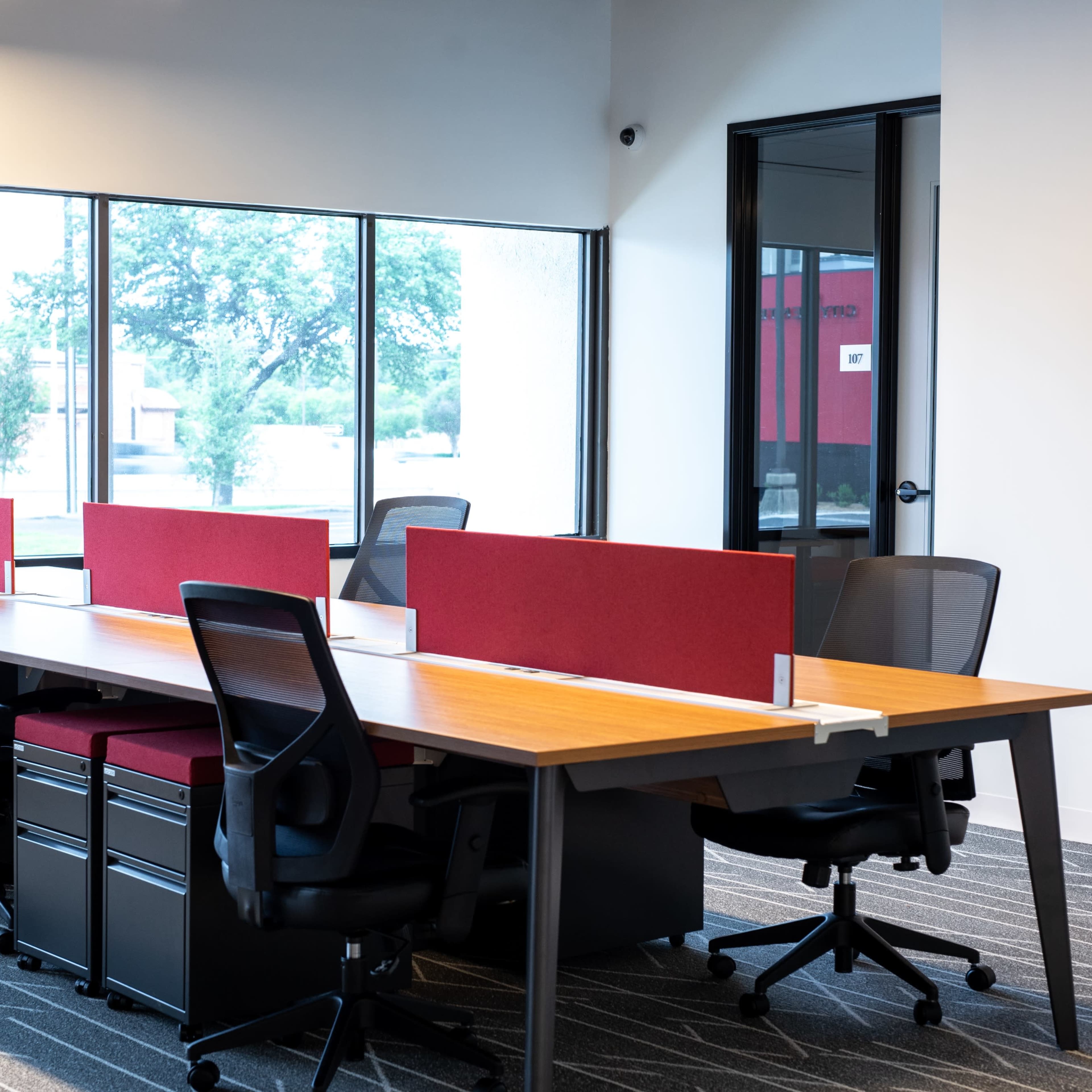 A modern office with a row of desks, each equipped with ergonomic chairs and partitions, beside large windows that overlook a landscaped area.