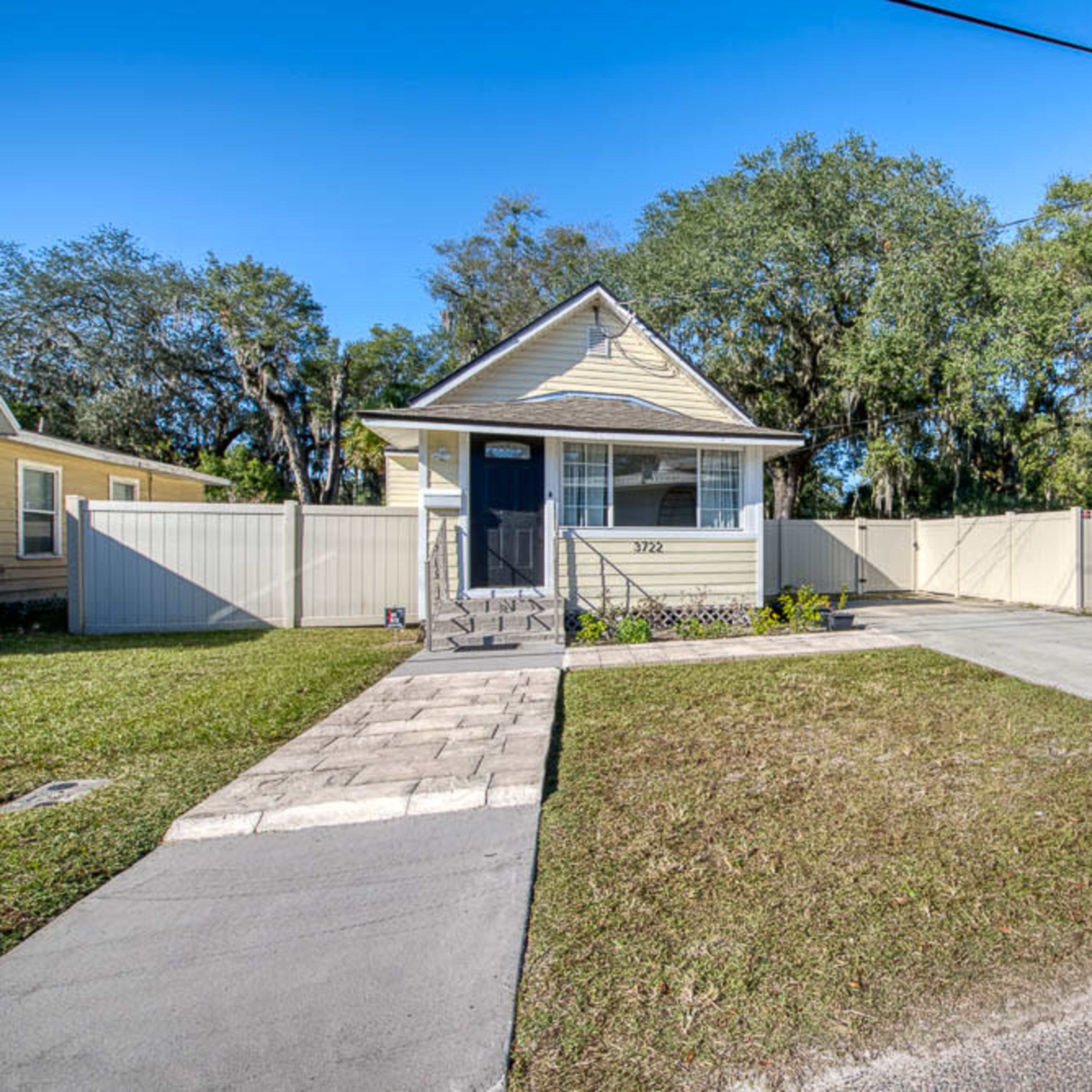 A small yellow house with a front porch, surrounded by a chain-link fence and a gravel driveway.