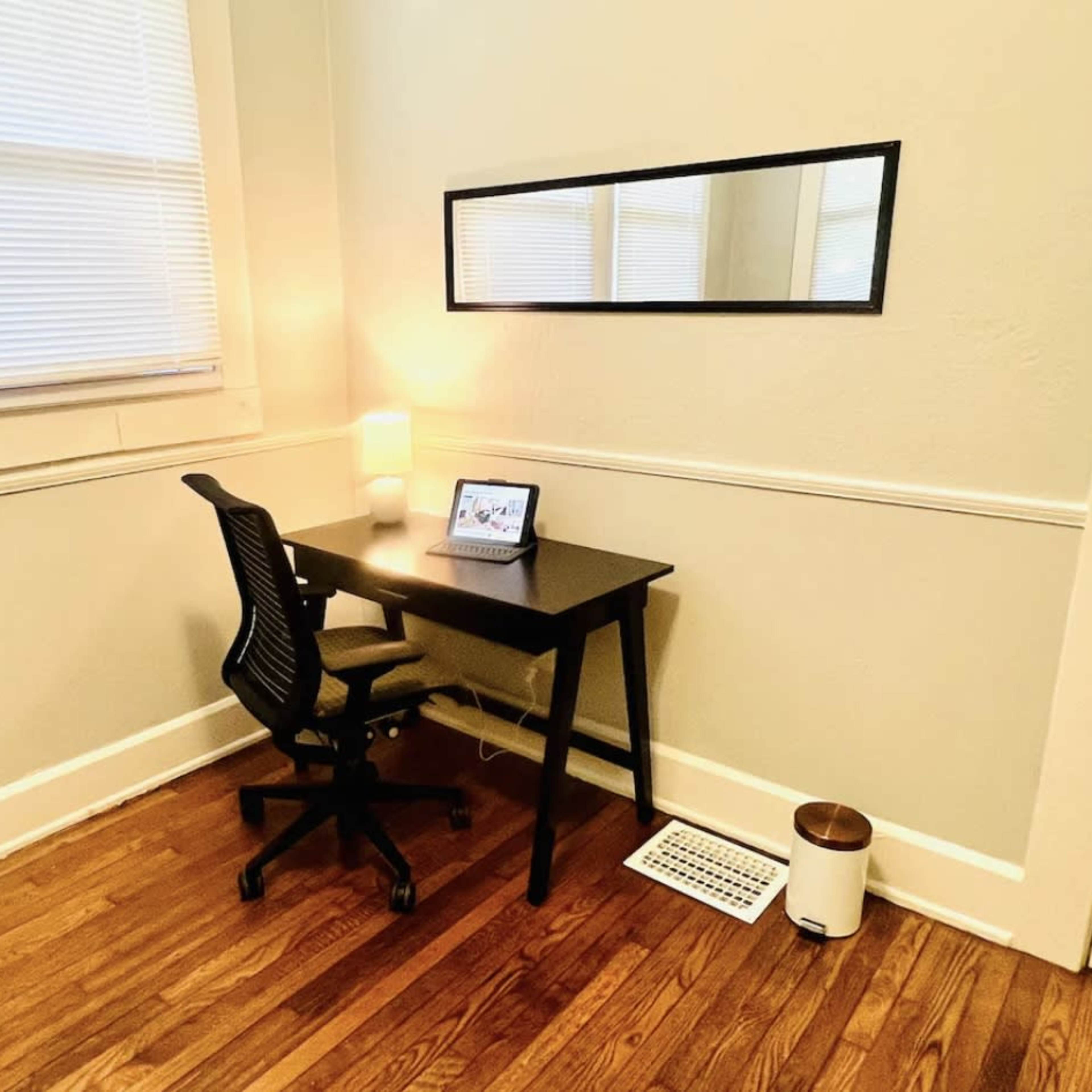 A small home office with a black desk, a rolling chair, a tablet on the desk, and a mirror mounted on the wall.