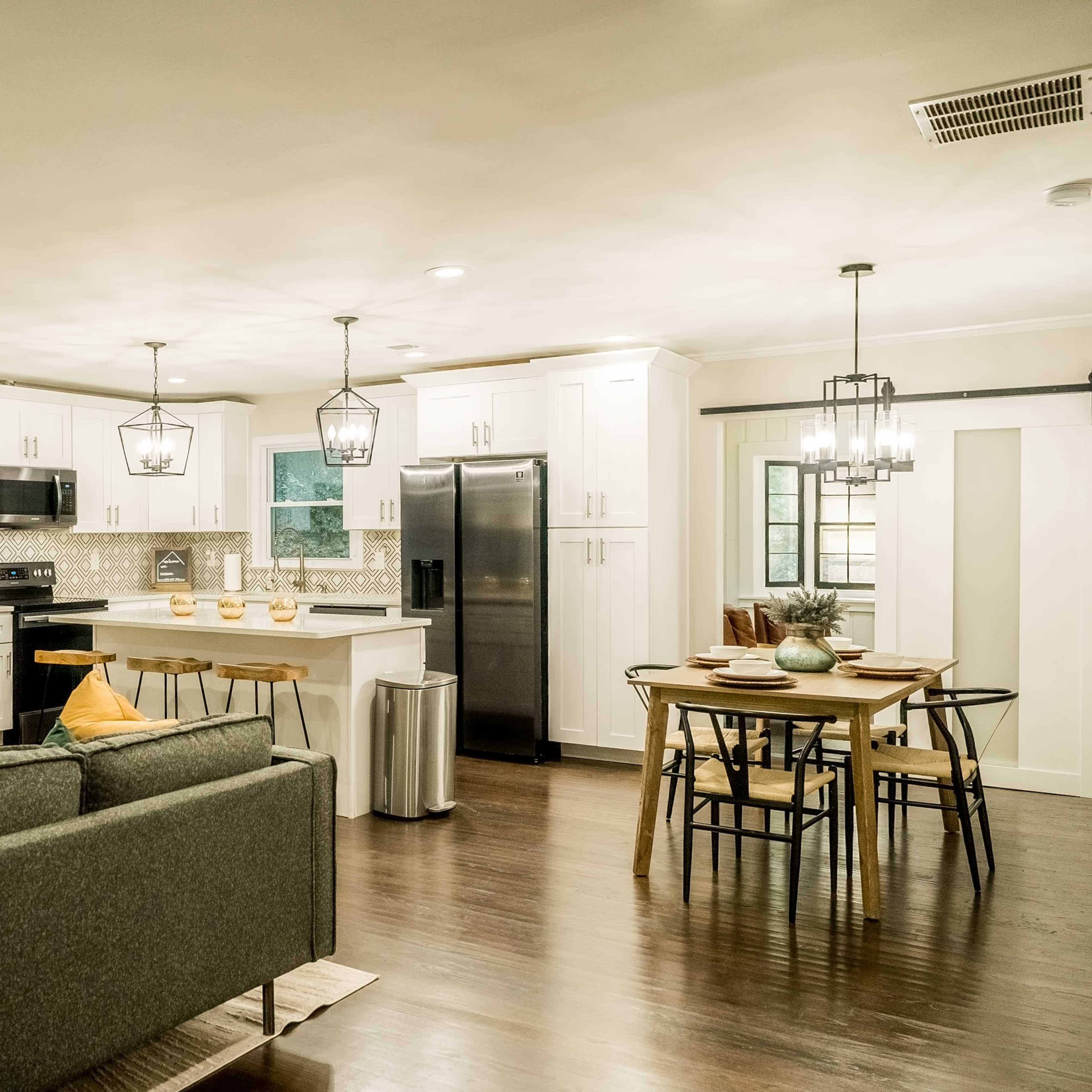 A modern kitchen with white cabinets and black appliances opens into a dining area featuring a table surrounded by chairs and a sofa in the foreground.