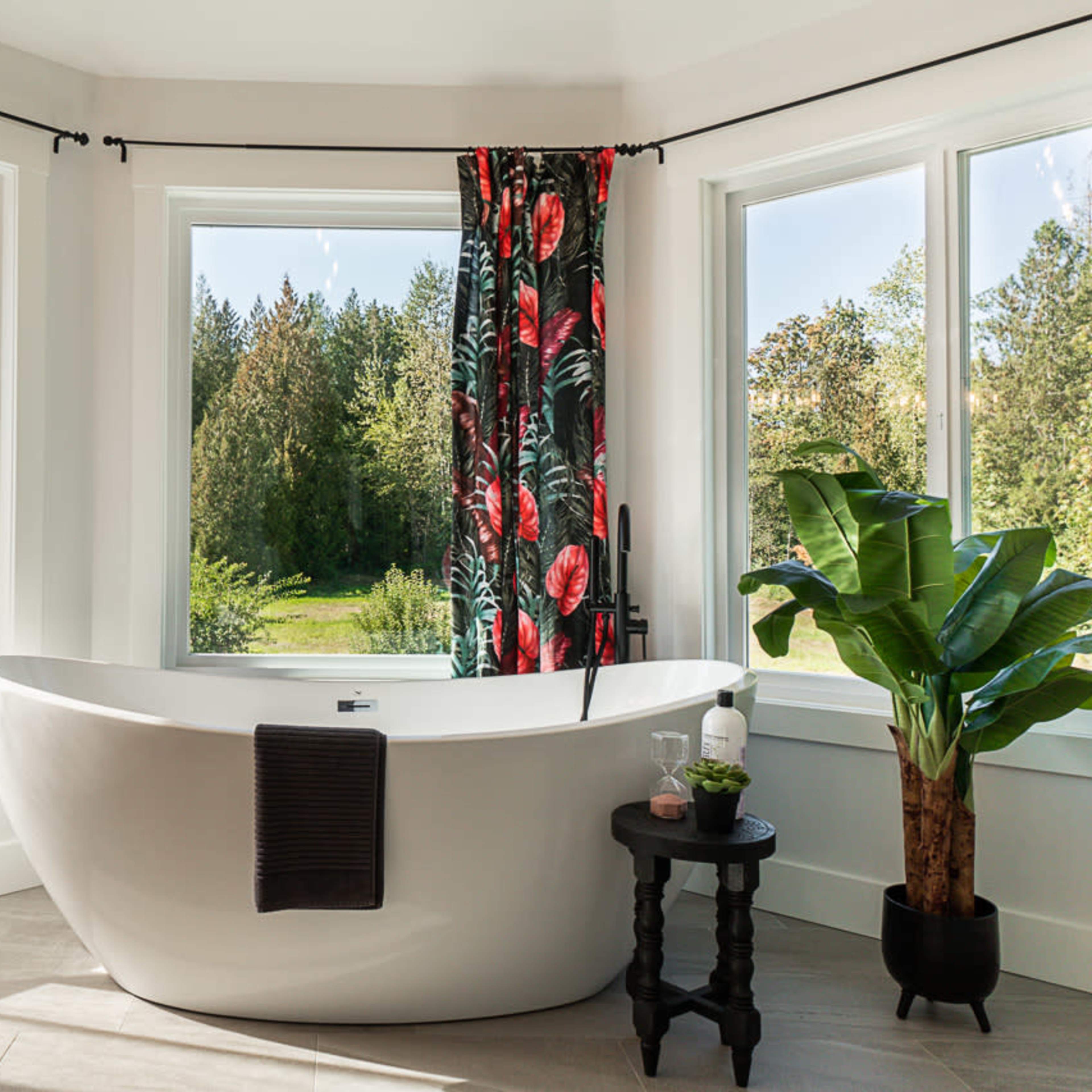 A freestanding white bathtub is positioned near large windows that offer a view of a green landscape, with sheer curtains and decorative plants enhancing the scene.