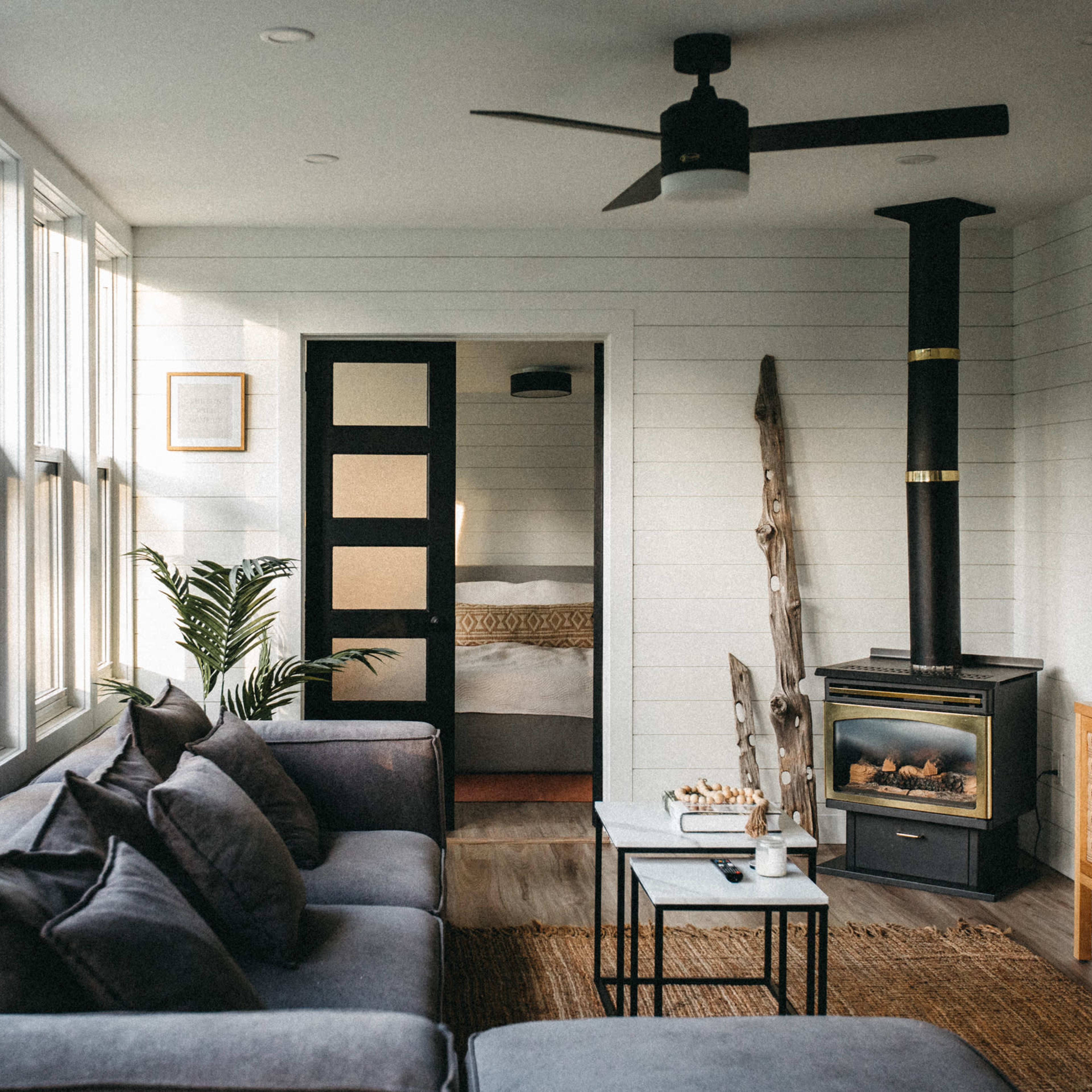 A modern living room features large windows, a gray sofa with decorative pillows, a black ceiling fan, and a wood stove, with a doorway leading to a bedroom in the background.