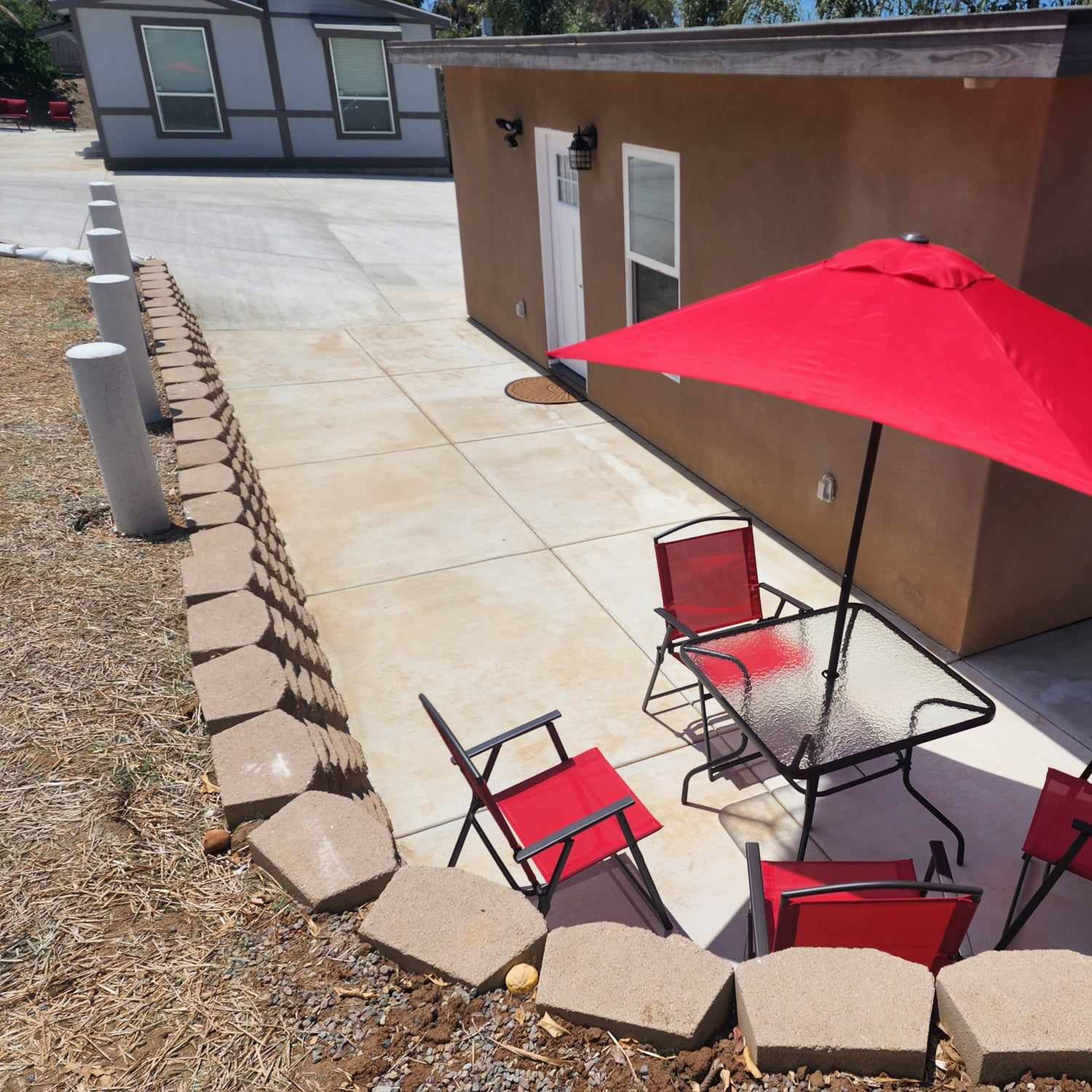 A patio area features a red umbrella over a black table with four red chairs, surrounded by a low stone wall near a building.