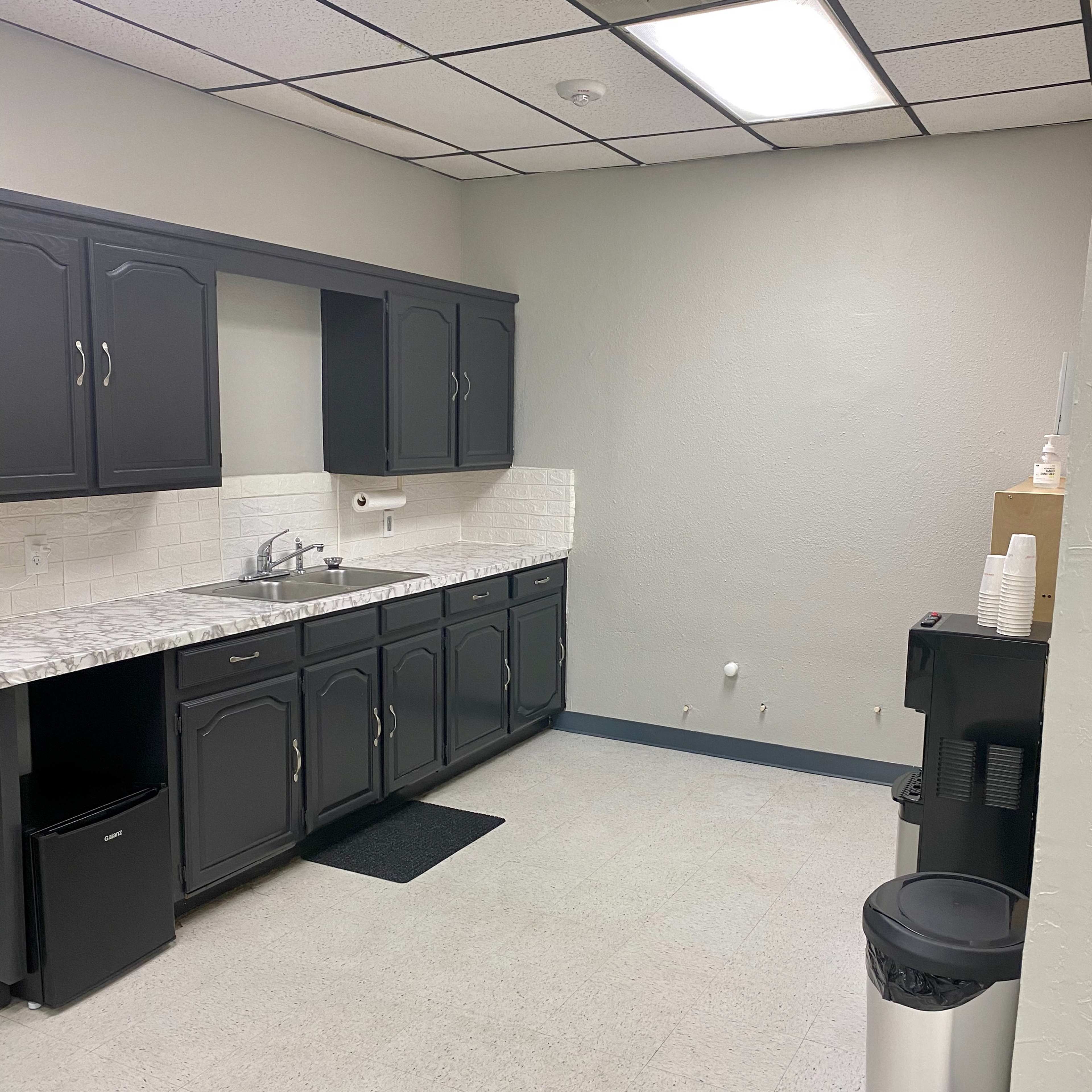 A clean, empty kitchenette with gray cabinets, a marble countertop, a sink, and a coffee maker beside a trash can.