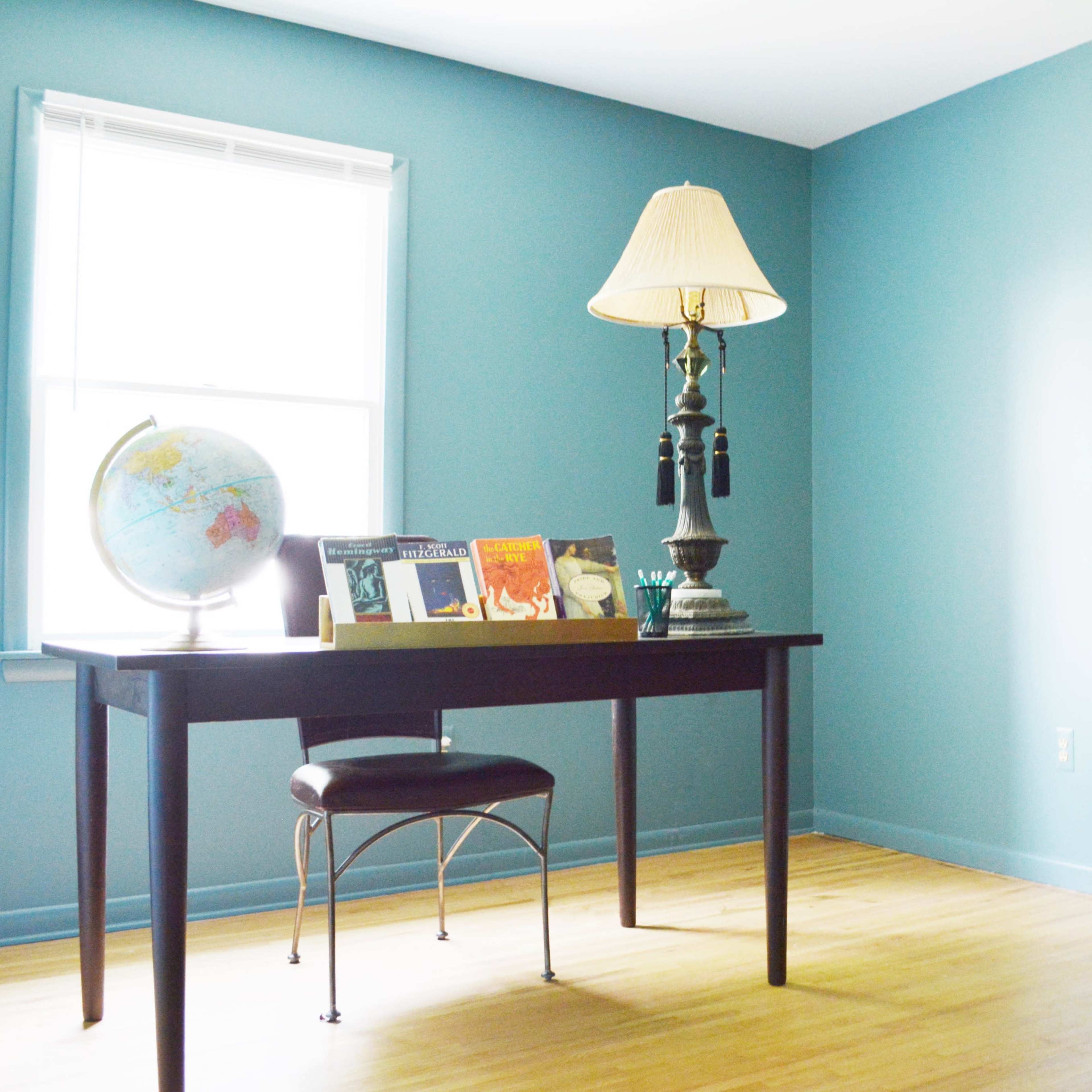 A small room features a wooden desk with a globe, books, a lamp, and a dining table with chairs against light blue walls.
