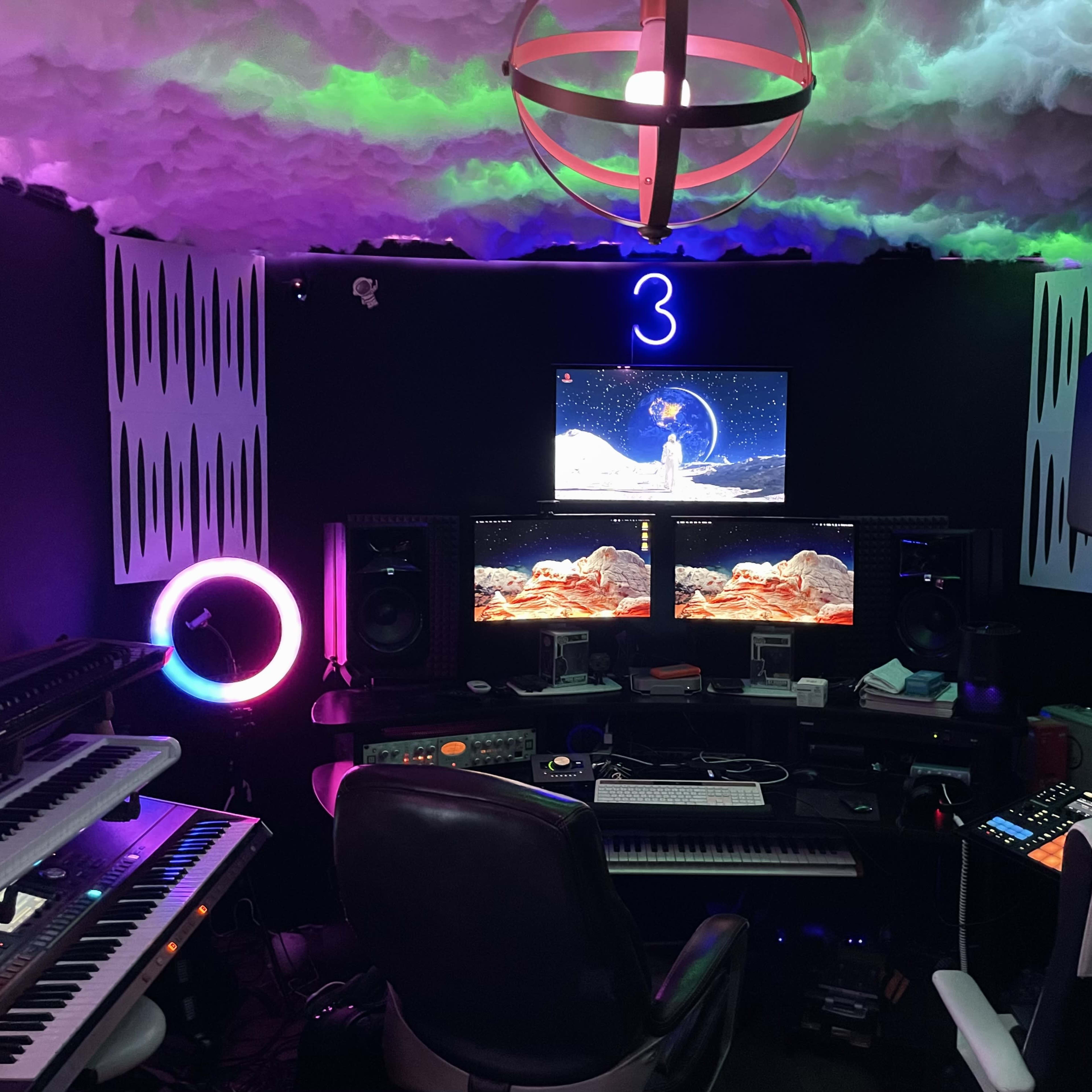 A home recording studio featuring multiple keyboards, two computer monitors displaying vibrant visuals, and colorful lighting on the ceiling.