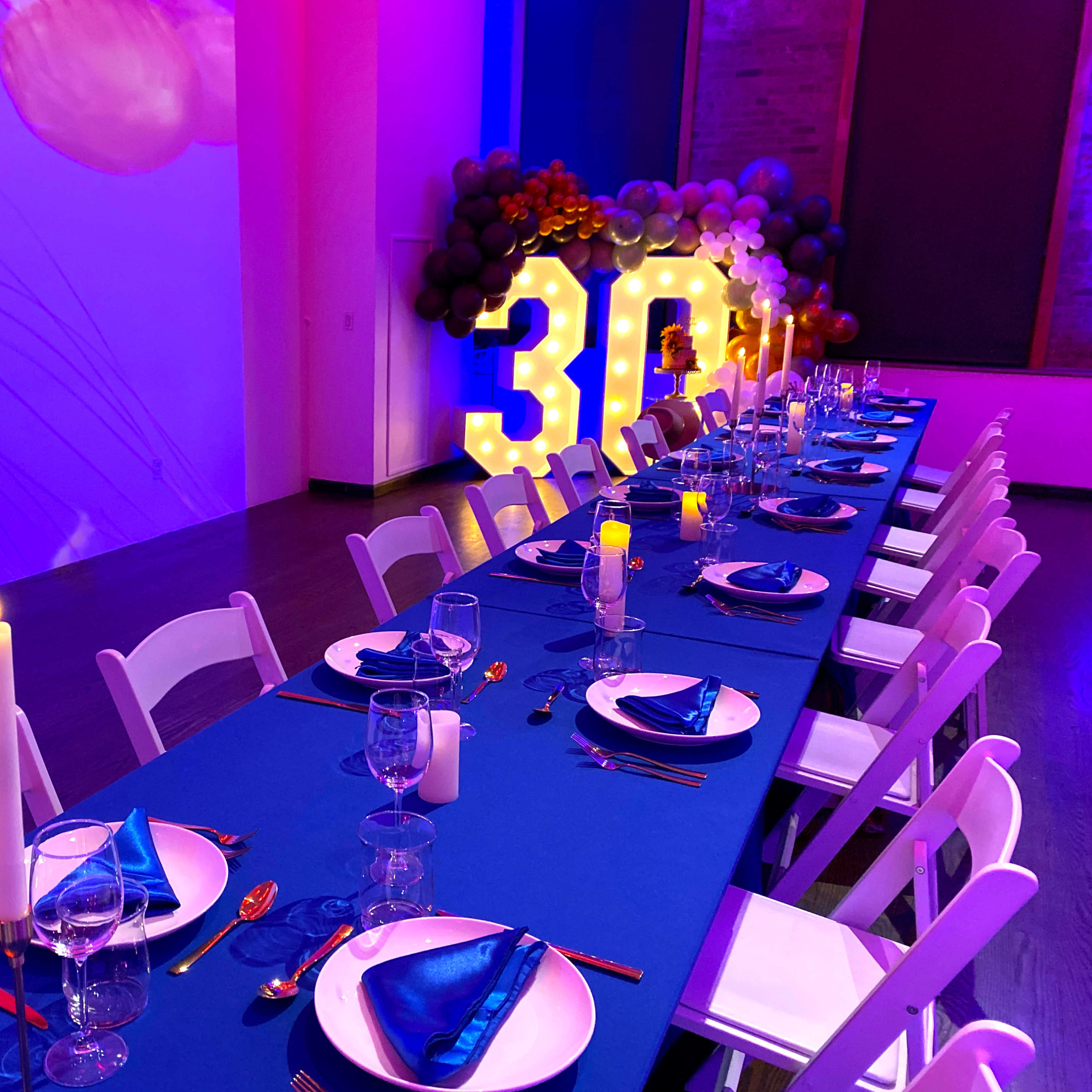 A long table is set for a celebration, adorned with blue tablecloth, elegant place settings, and illuminated candles, while large decorative lights reading "30" are showcased in the background.