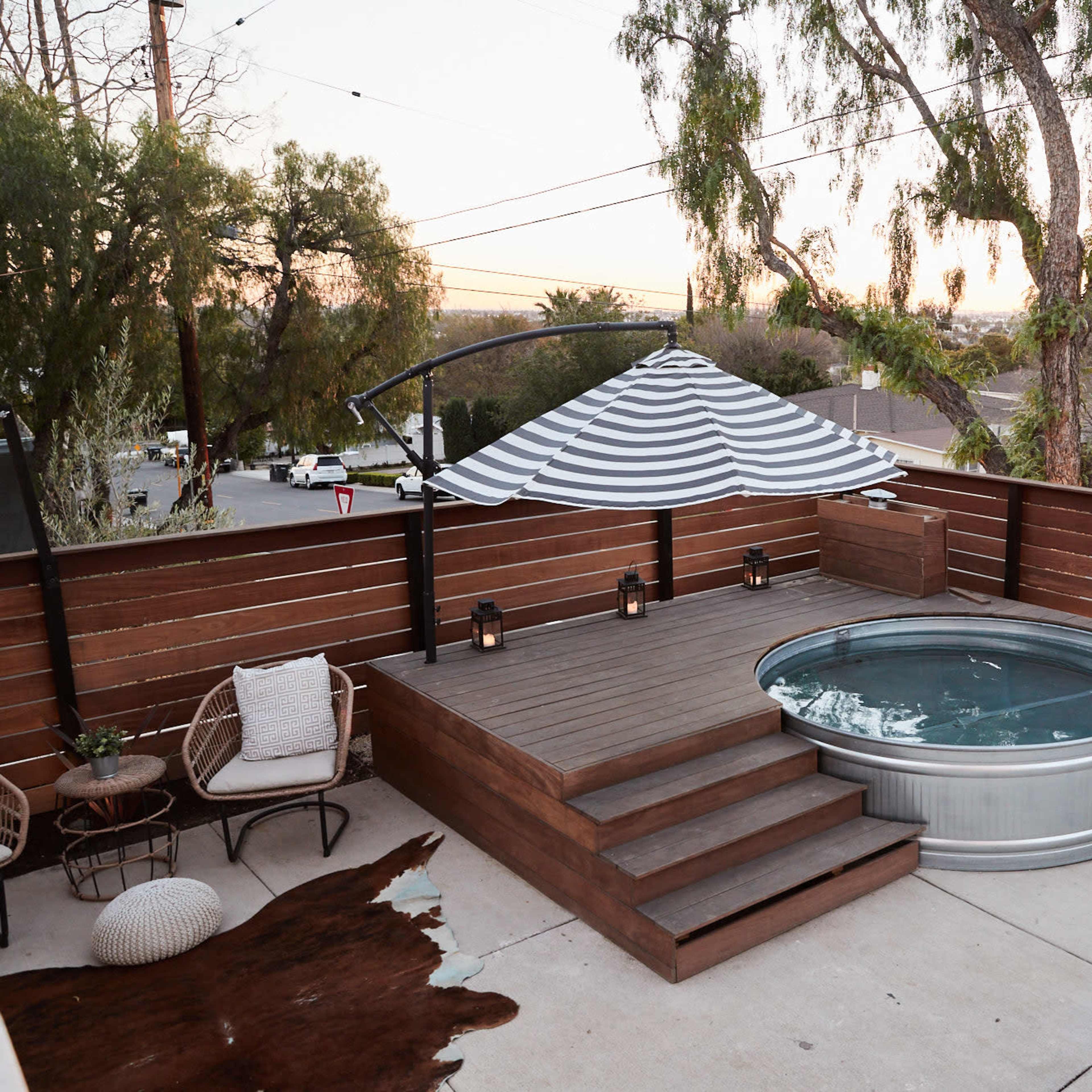 A wooden deck features a round hot tub with steps and a striped canopy, surrounded by a fenced area and sunset views.