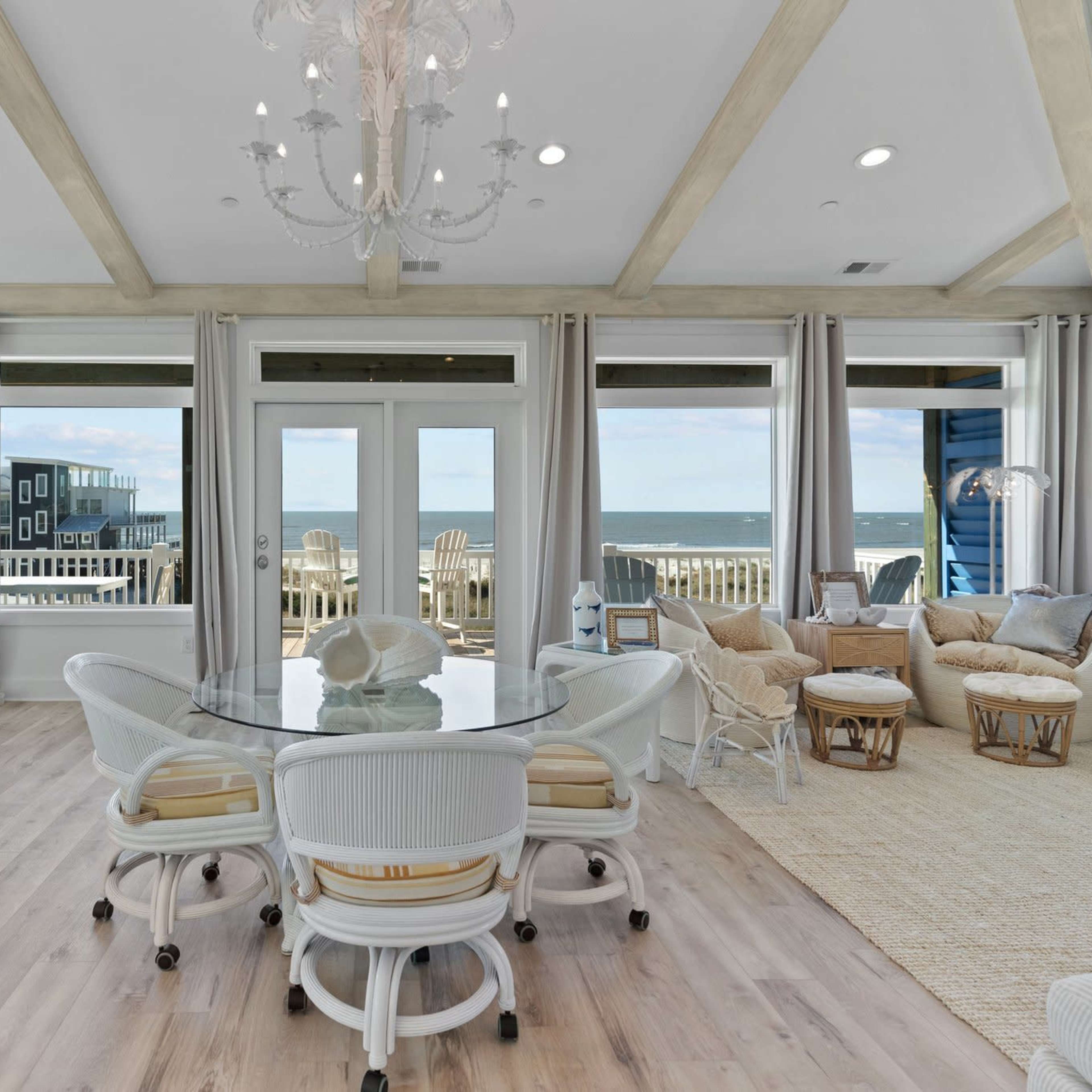 A bright, airy living space with large windows overlooking the ocean, a round dining table surrounded by white chairs, and cozy seating areas.