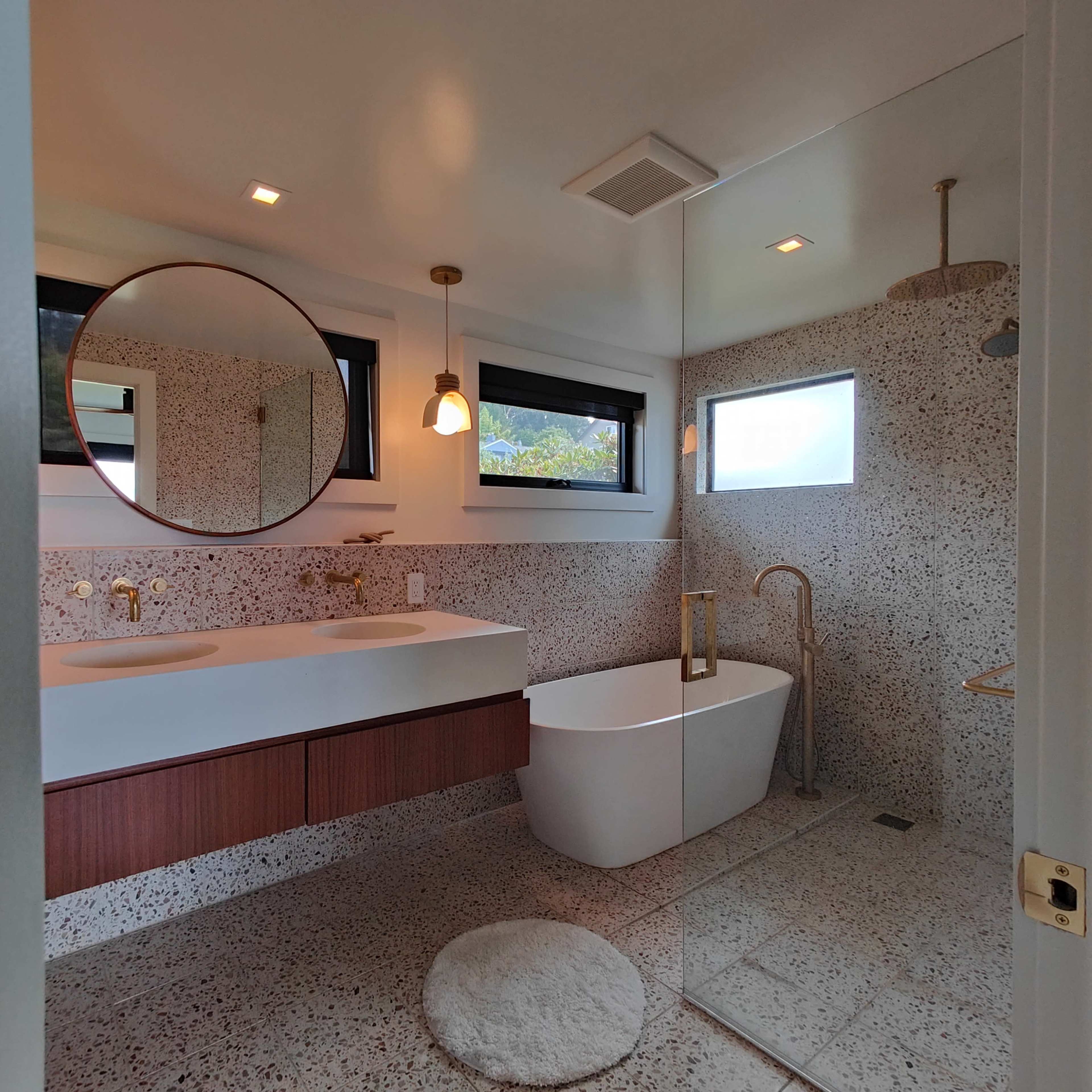 A modern bathroom with a double sink vanity, a freestanding bathtub, and a textured wall of large speckled tiles.