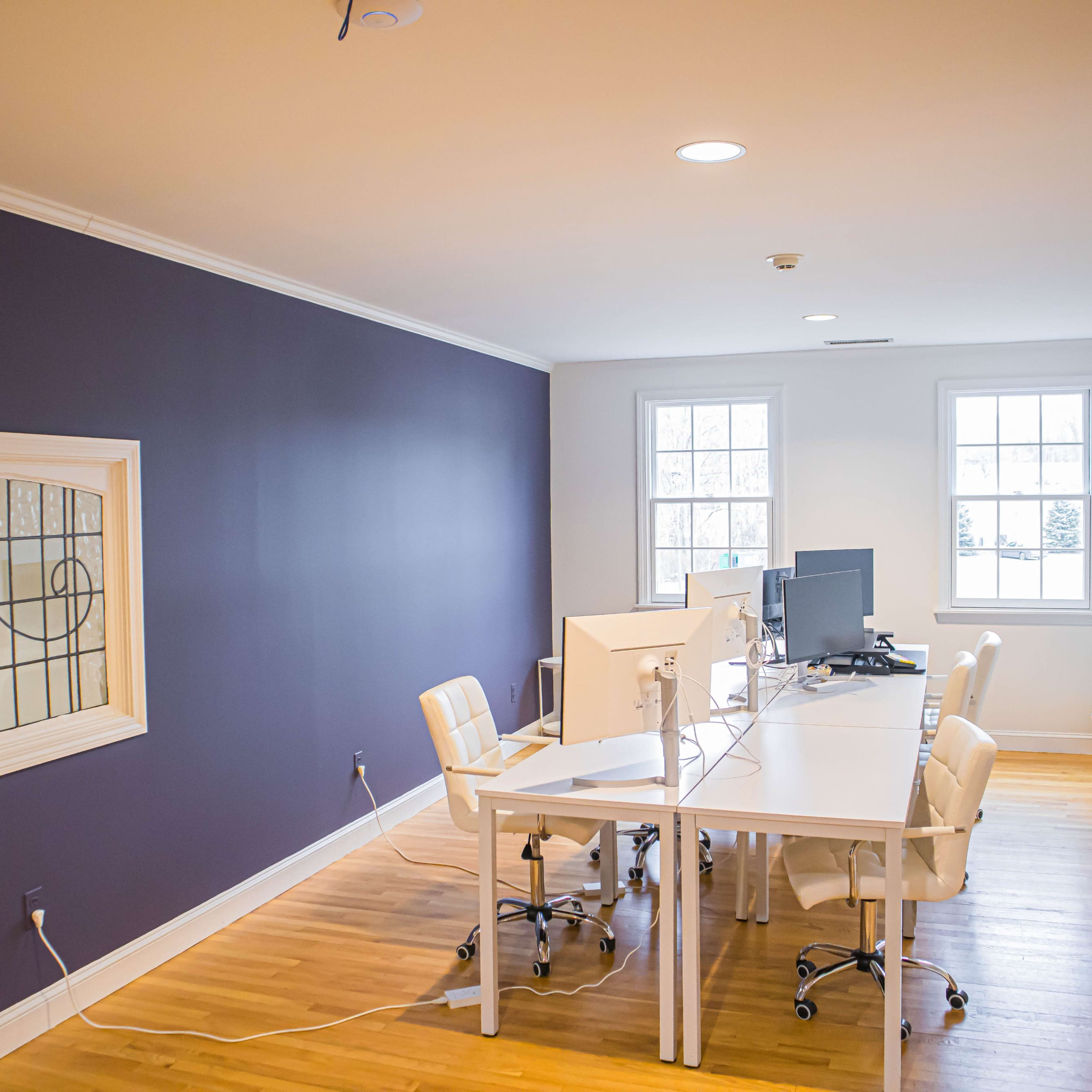 A minimalist office space with a white desk, two computer monitors, and white chairs against a purple wall.