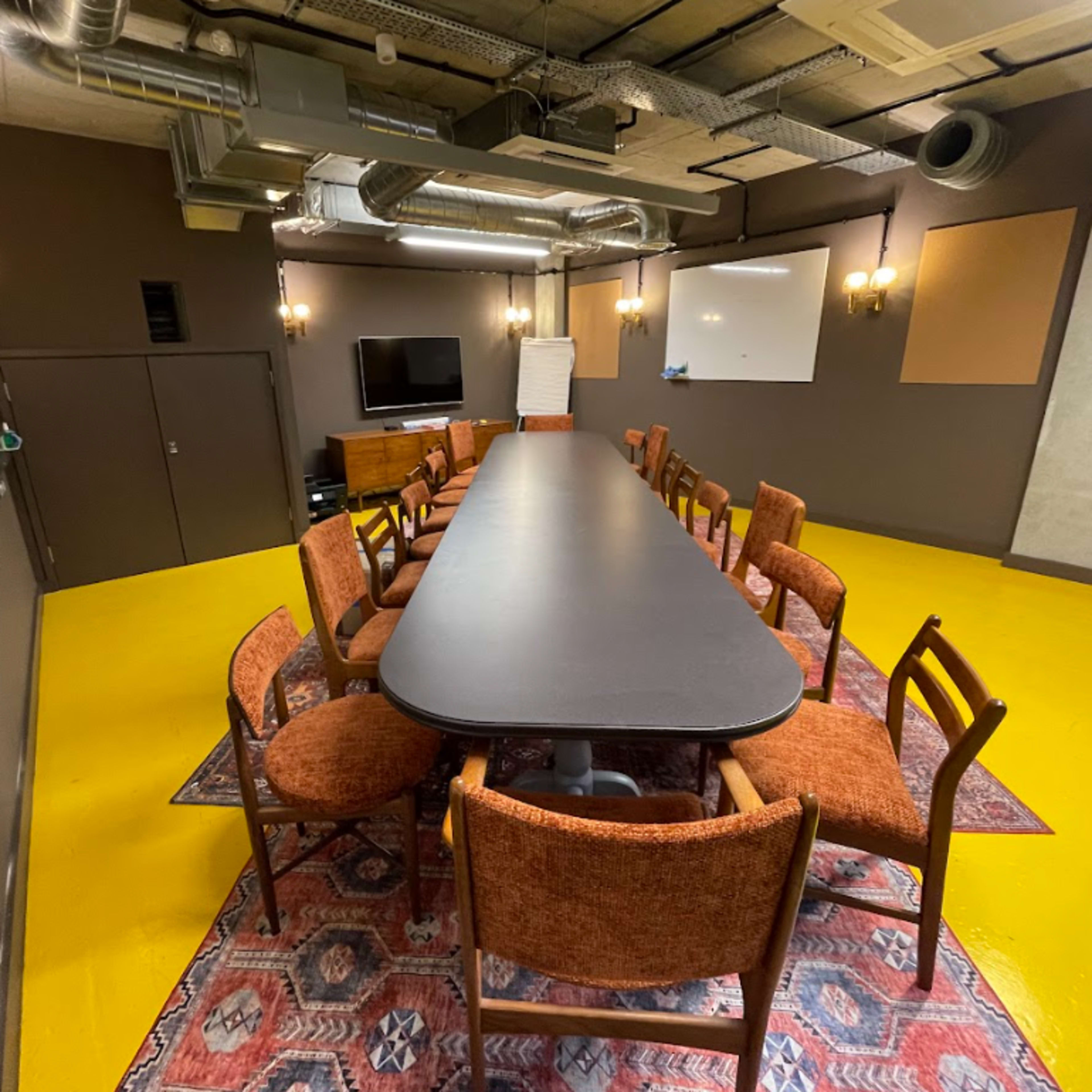 A conference room features a long black table surrounded by wooden chairs on a yellow floor, with a television and whiteboard mounted on the walls.