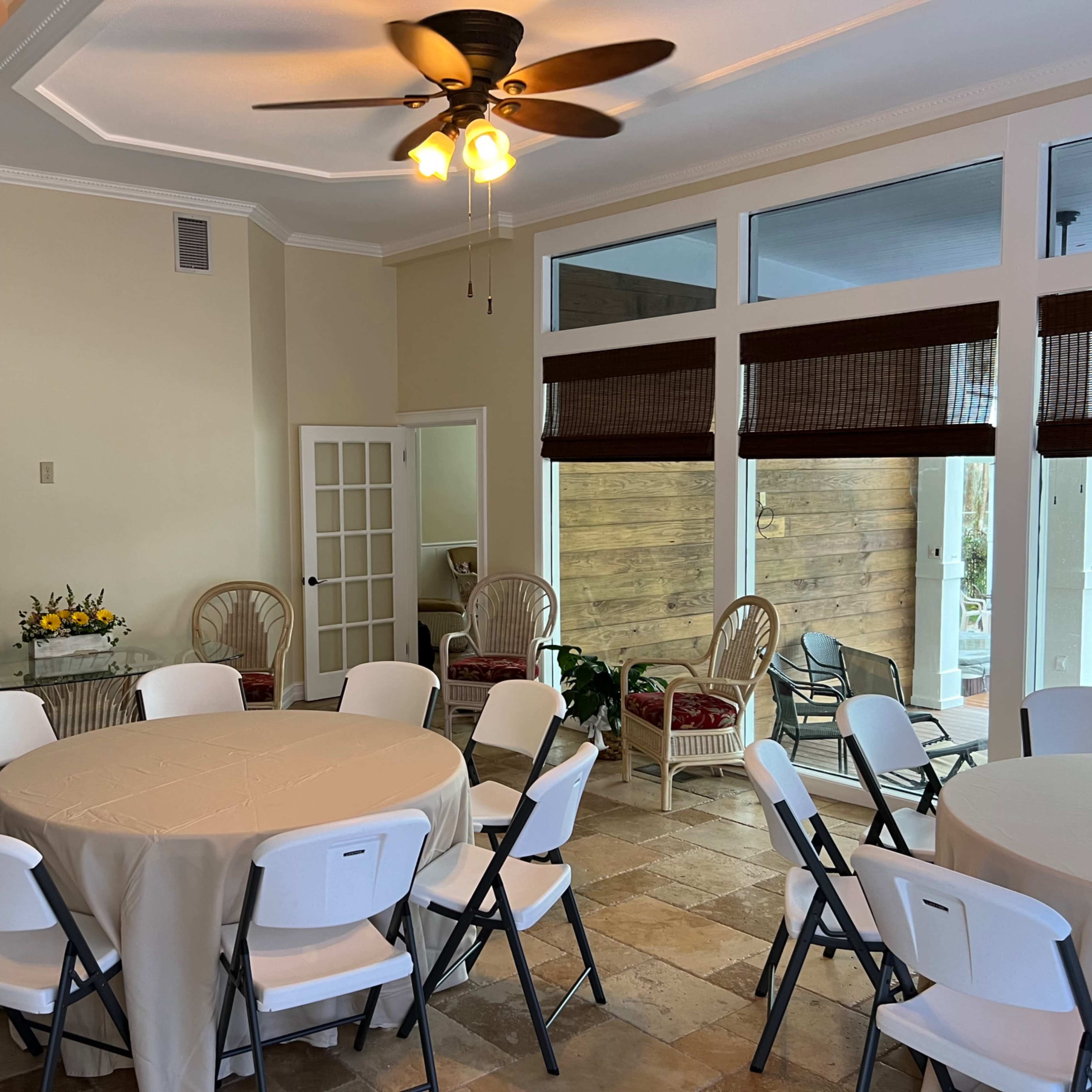 A spacious room set up with circular tables covered in white tablecloths, surrounded by folding chairs, with large windows and a small seating area in the background.