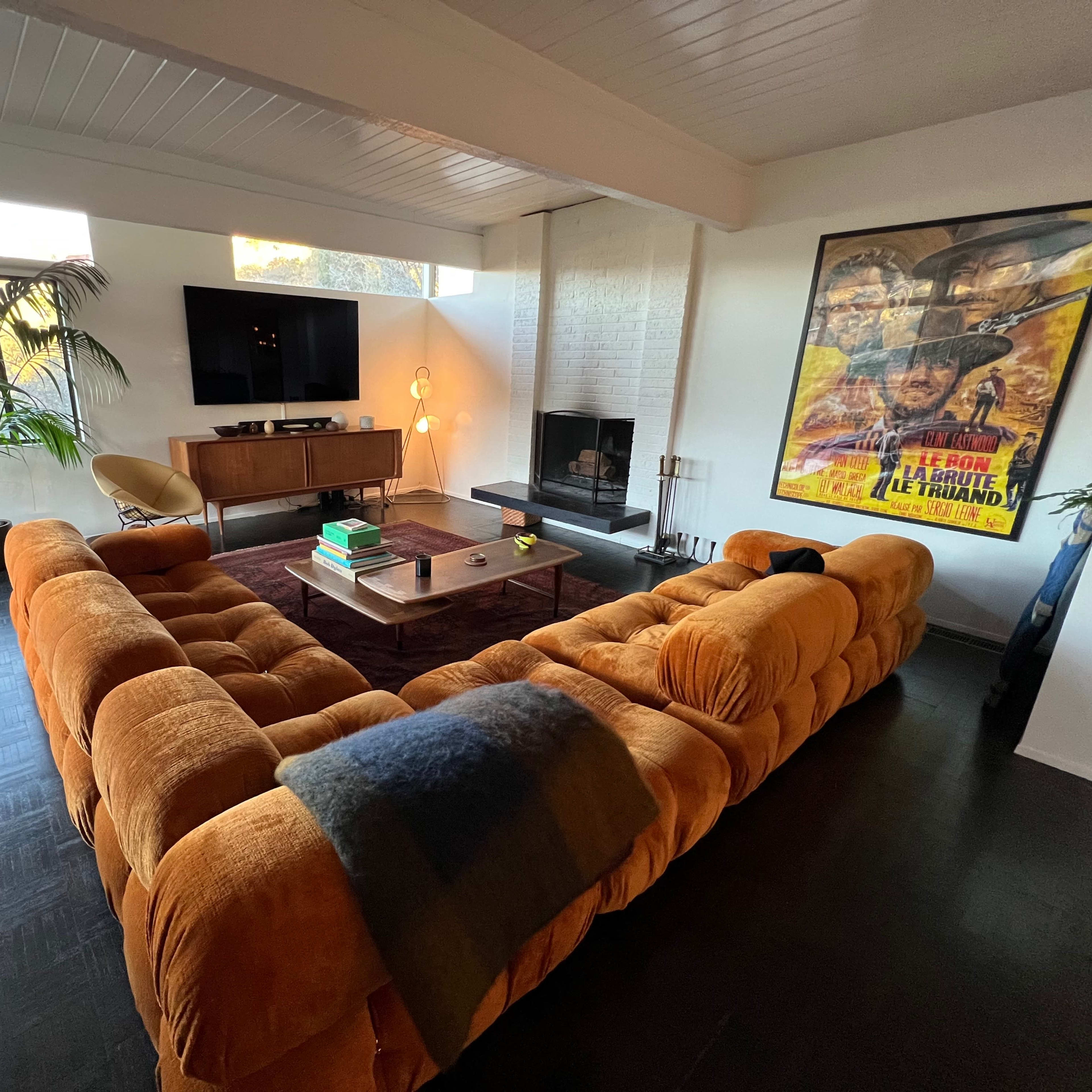 A modern living room featuring a large, orange sectional sofa, a coffee table, a television mounted on the wall, and a fireplace, with plants and artwork enhancing the space.