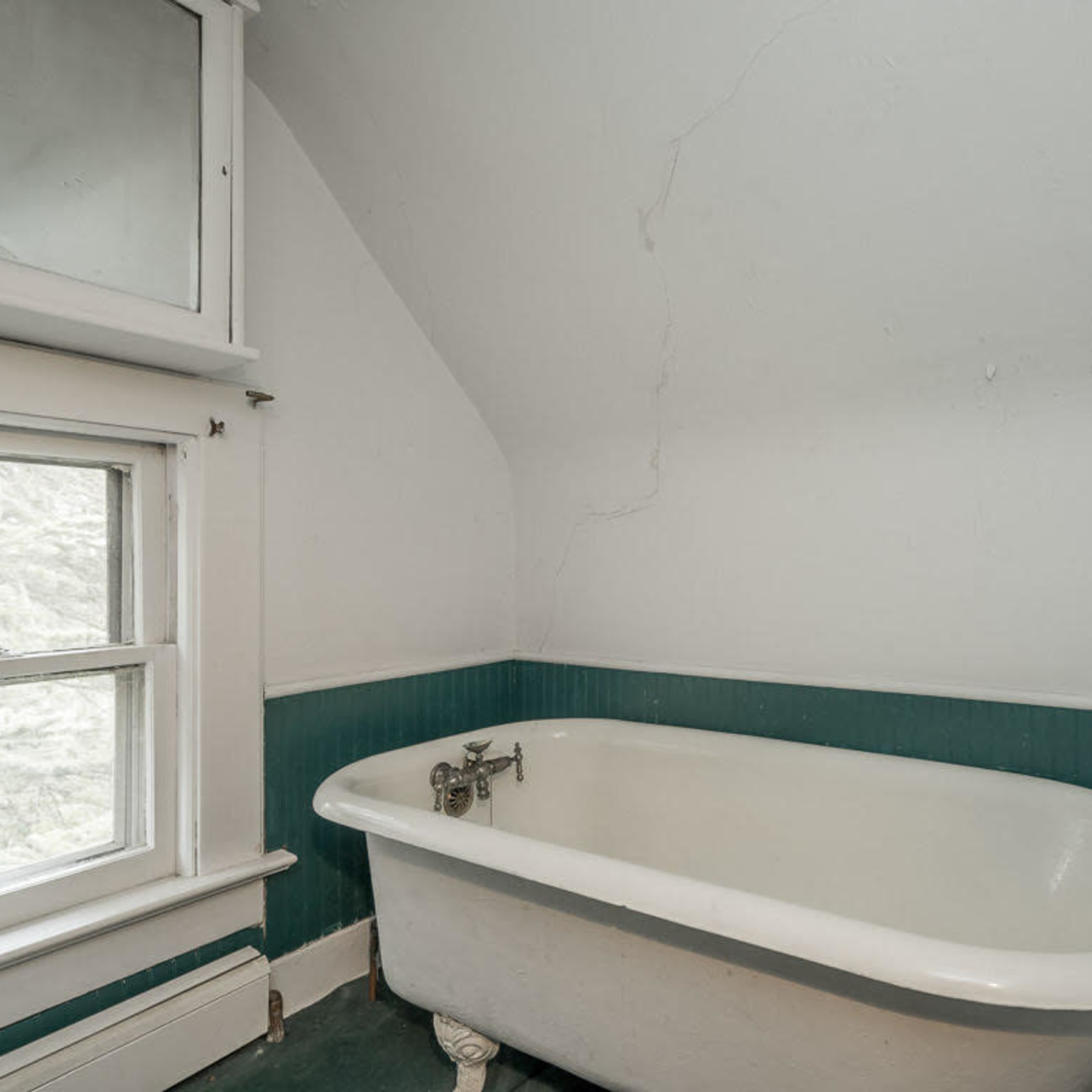A freestanding clawfoot bathtub is positioned in a small, angled bathroom with a window and beadboard paneling.