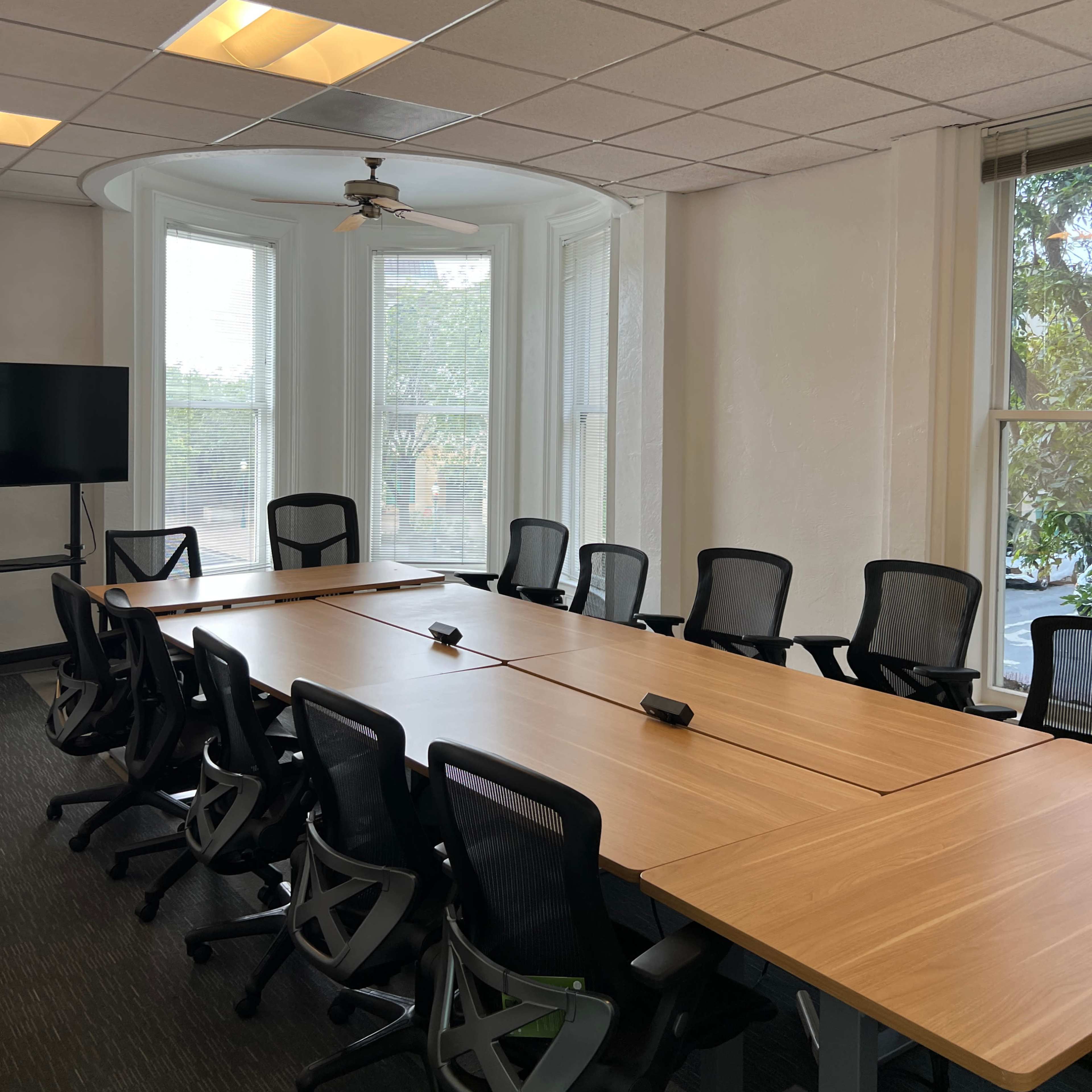 A conference room features a long wooden table surrounded by ergonomic chairs, with windows overlooking greenery and a television screen mounted on the wall.
