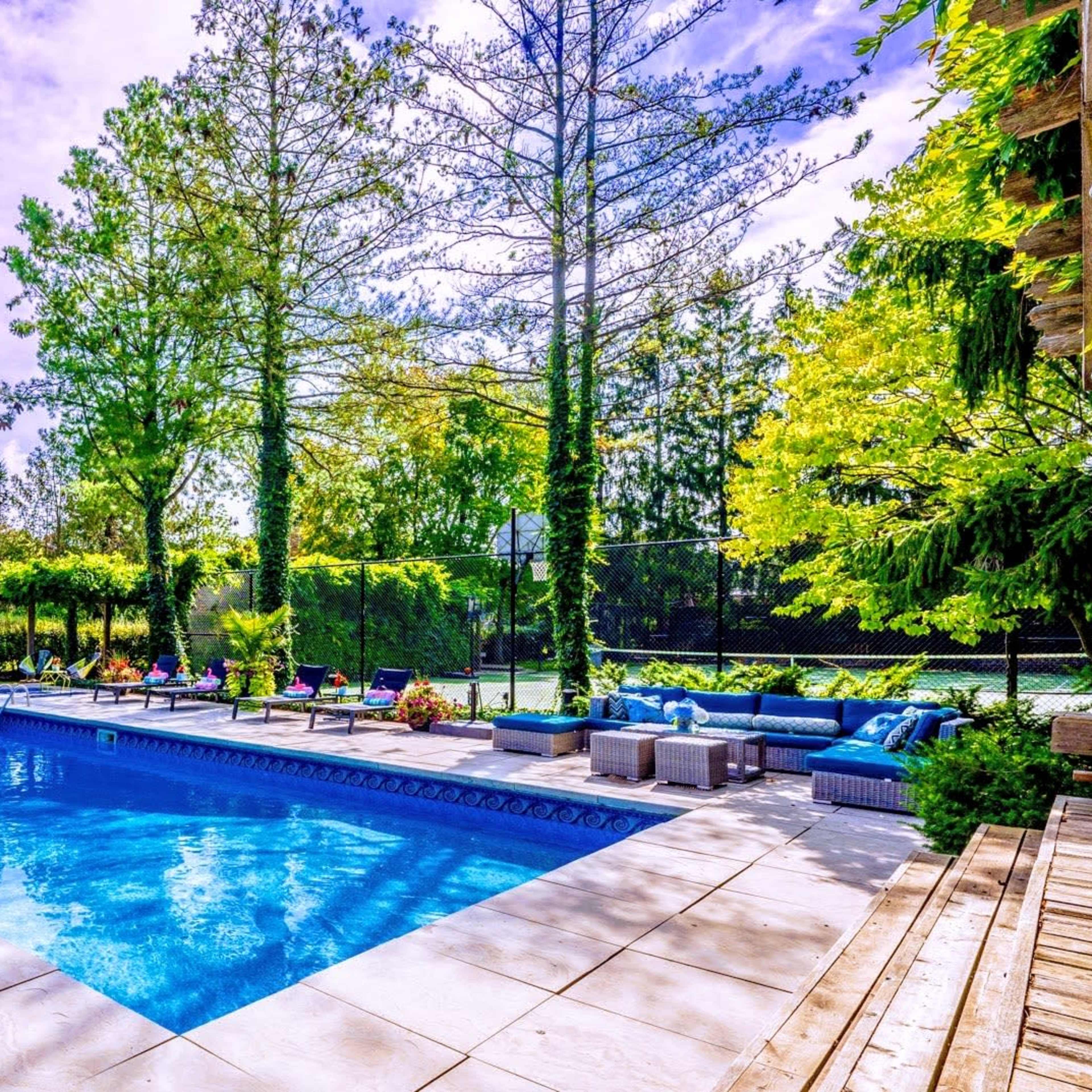A backyard swimming pool surrounded by green trees and lounge furniture, with colorful floaties in the water.
