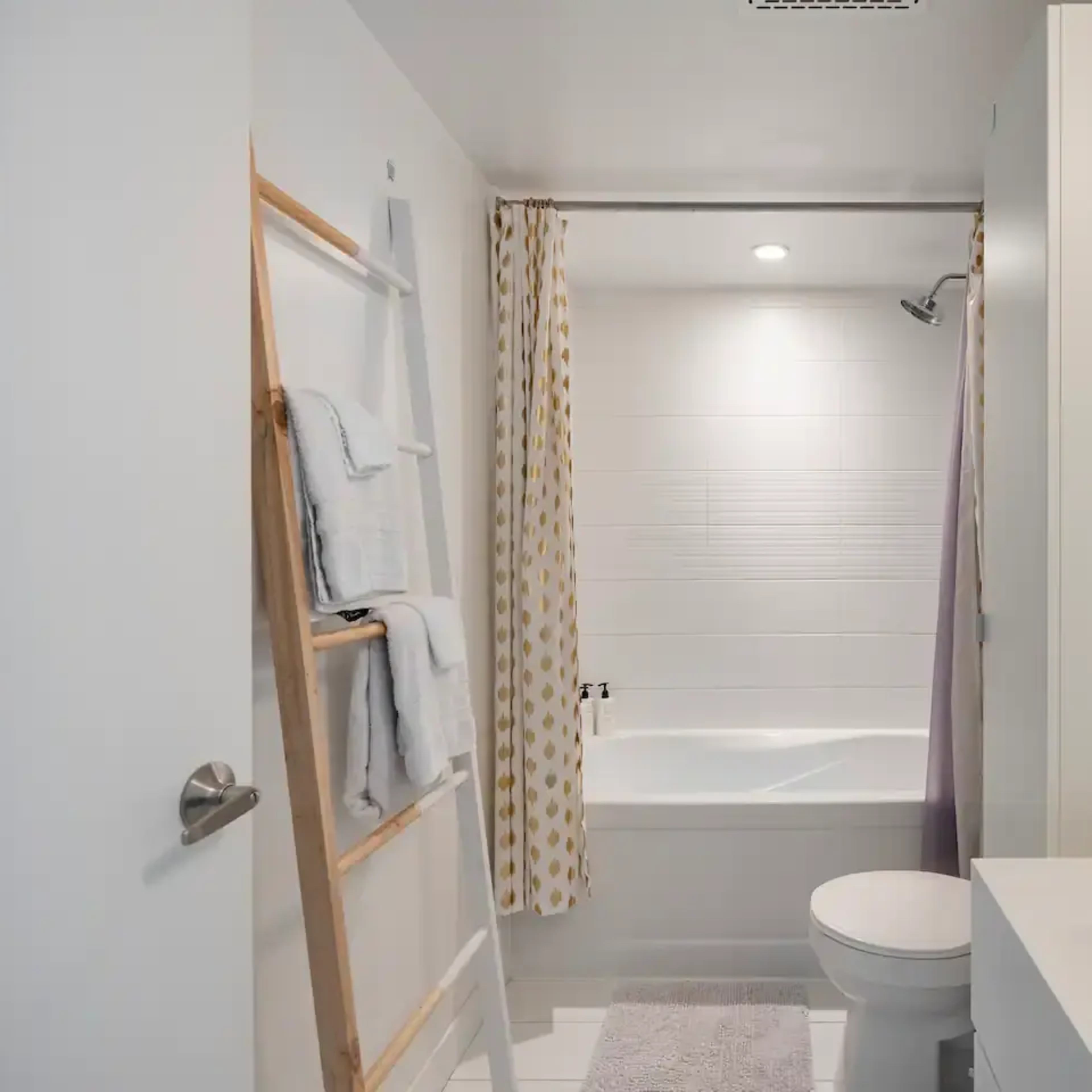 A modern bathroom featuring a bathtub, a toilet, a wooden ladder with towels, and a sink area with toiletries arranged on a countertop.