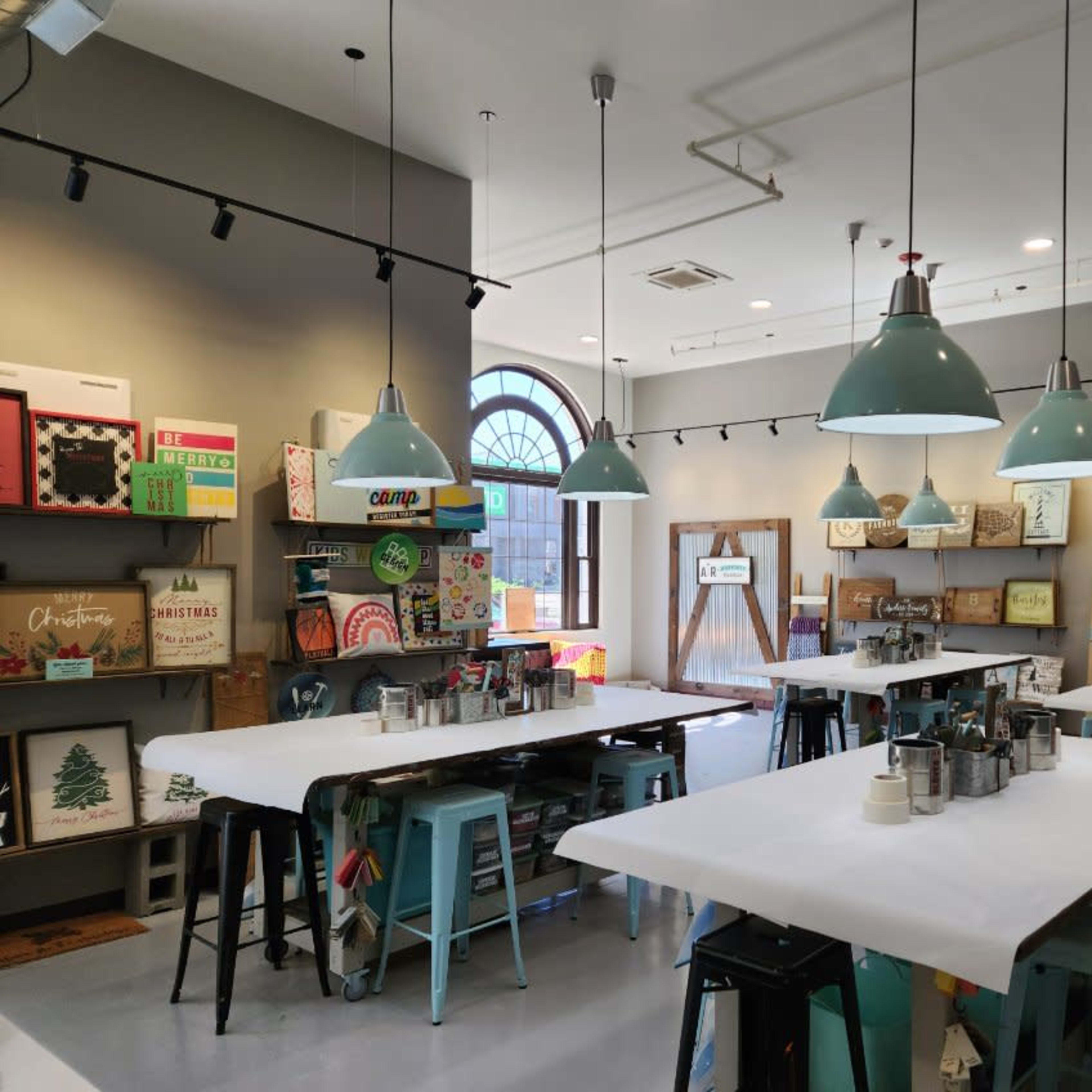 A spacious crafting studio with several tables, teal pendant lights, and shelves displaying various art supplies and decorative items.