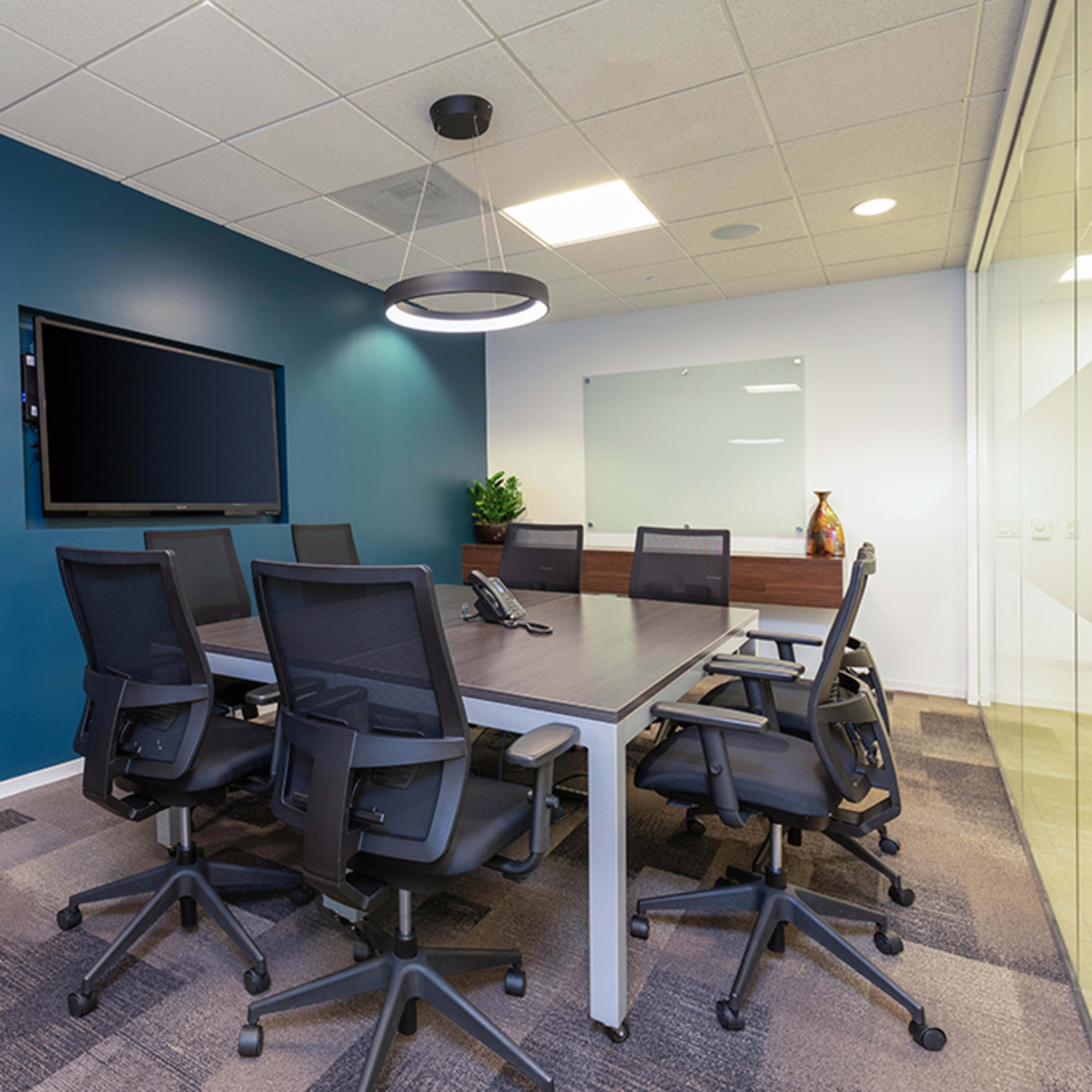 A modern conference room with a large table, black chairs, a wall-mounted television, and a glass-paneled wall.