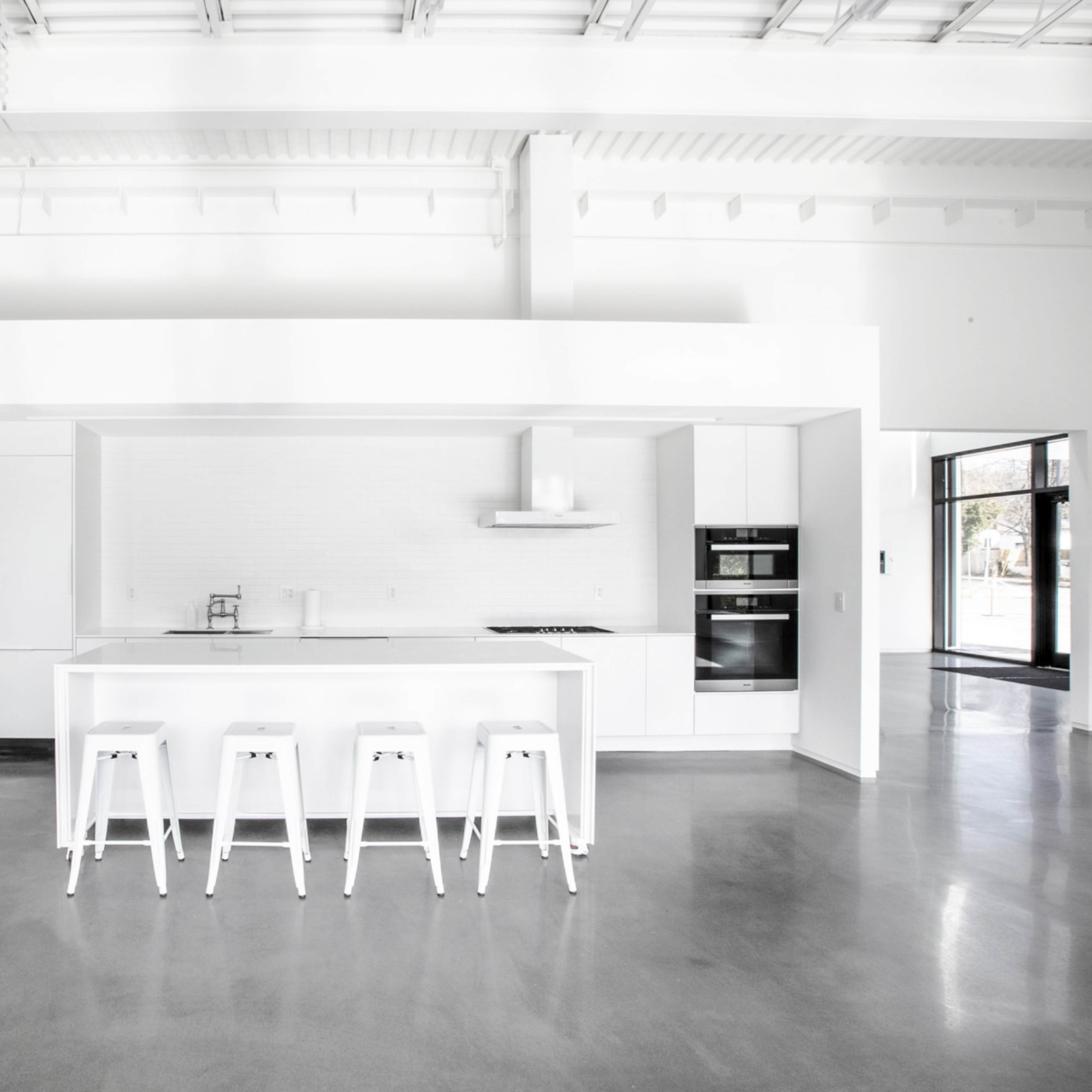 A modern kitchen with a minimalist design, featuring a white island and four white stools, set in a spacious, light-filled area with concrete floors.