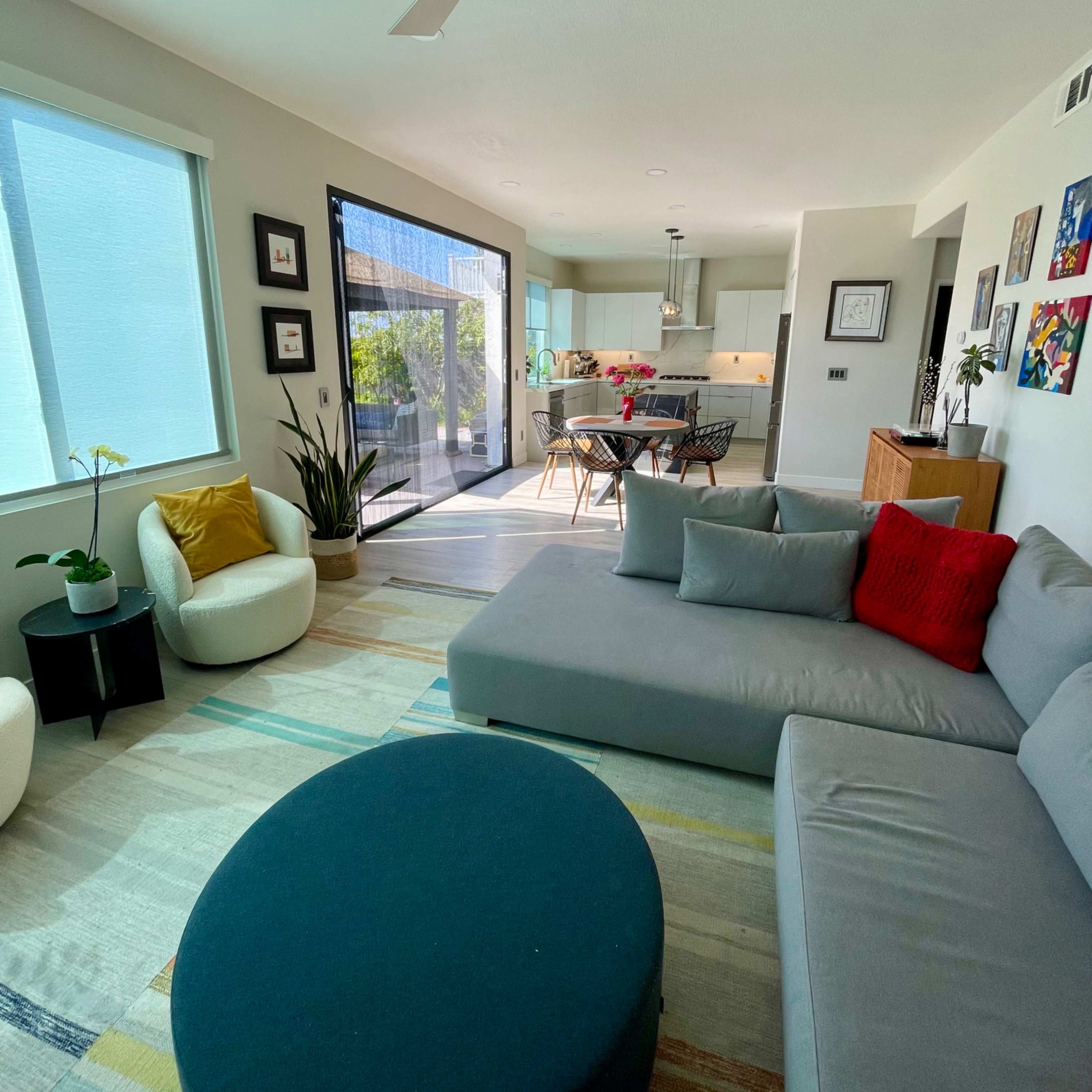 A modern living room with a sectional sofa, an ottoman, and a dining area in an open-concept space, featuring large windows that provide natural light.
