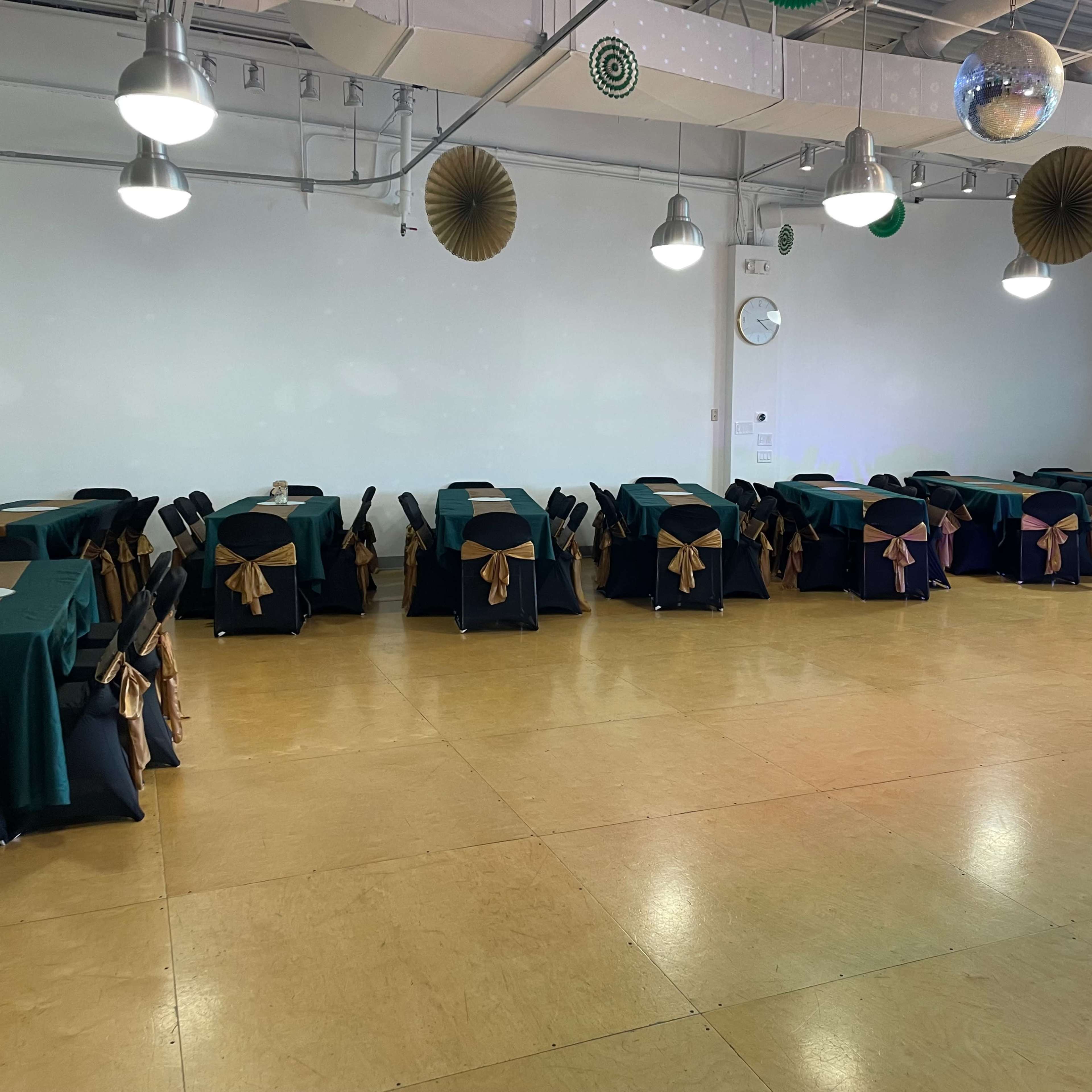 A large, empty banquet hall features rows of tables covered with teal and black tablecloths, adorned with gold sashes.