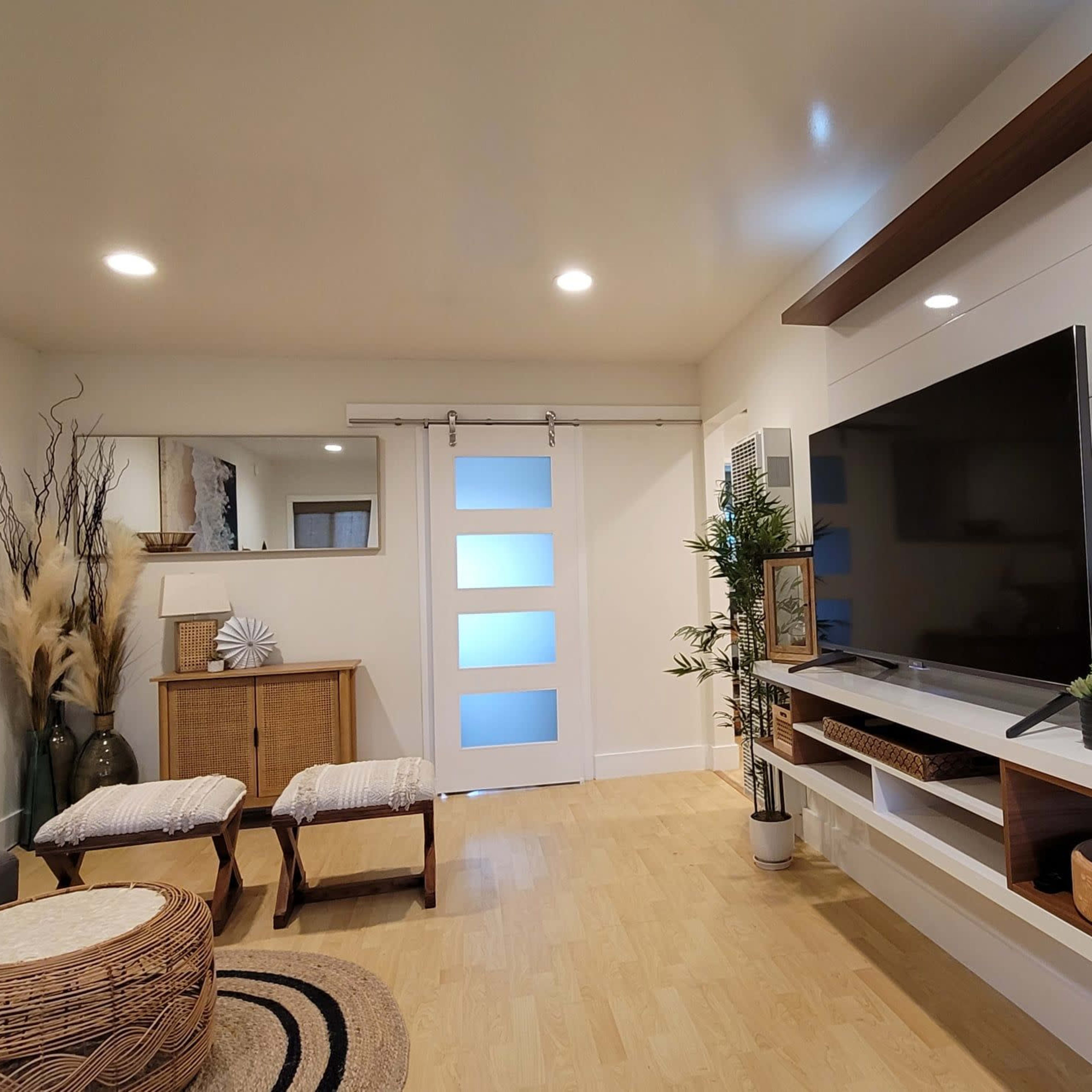 The living room features a contemporary design with a wall-mounted television, a sliding glass door, and natural decor elements like wicker baskets and potted plants.