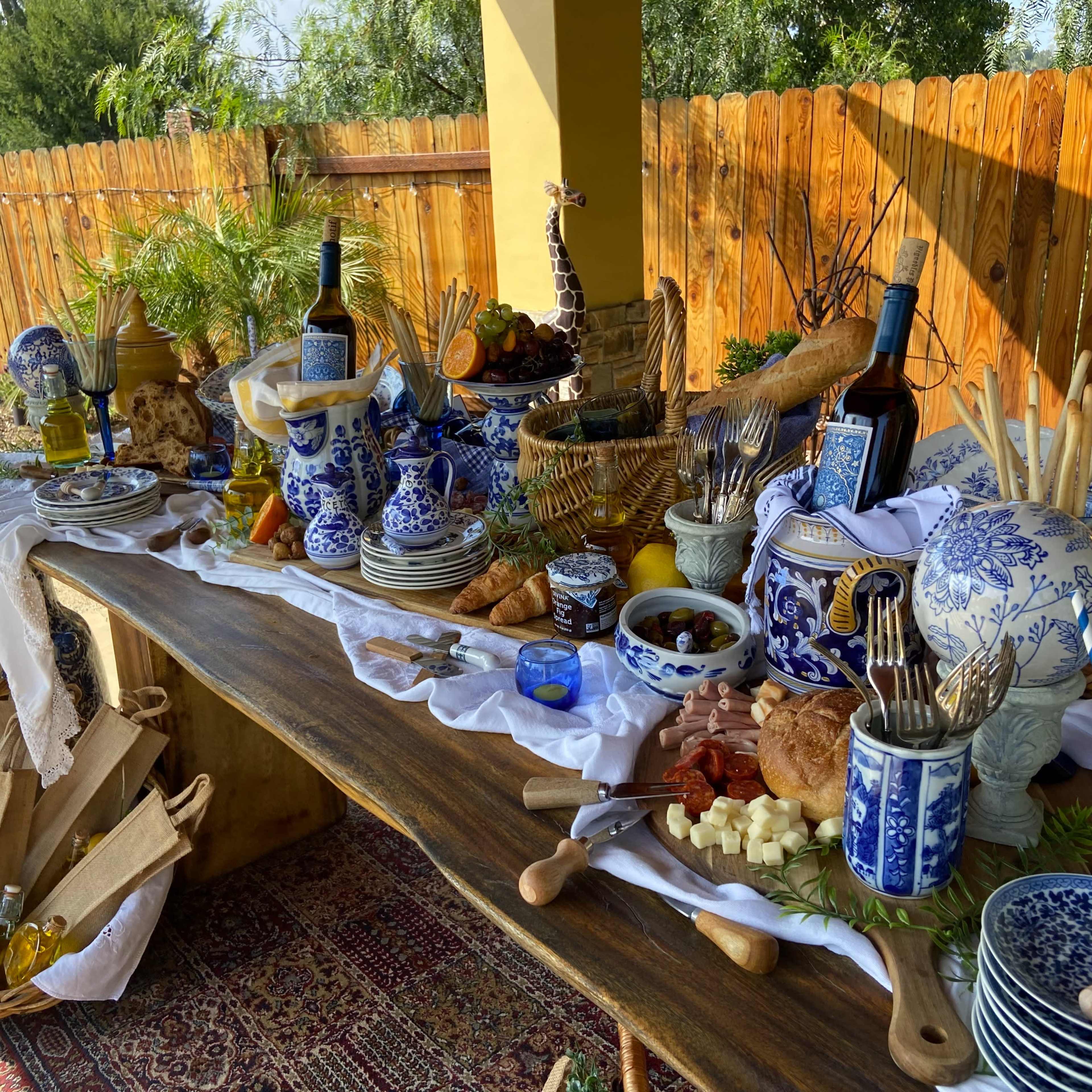 A wooden table is set with a variety of dishes, utensils, and decorative items, showcasing a spread of food and beverages accompanied by blue and white ceramic ware.