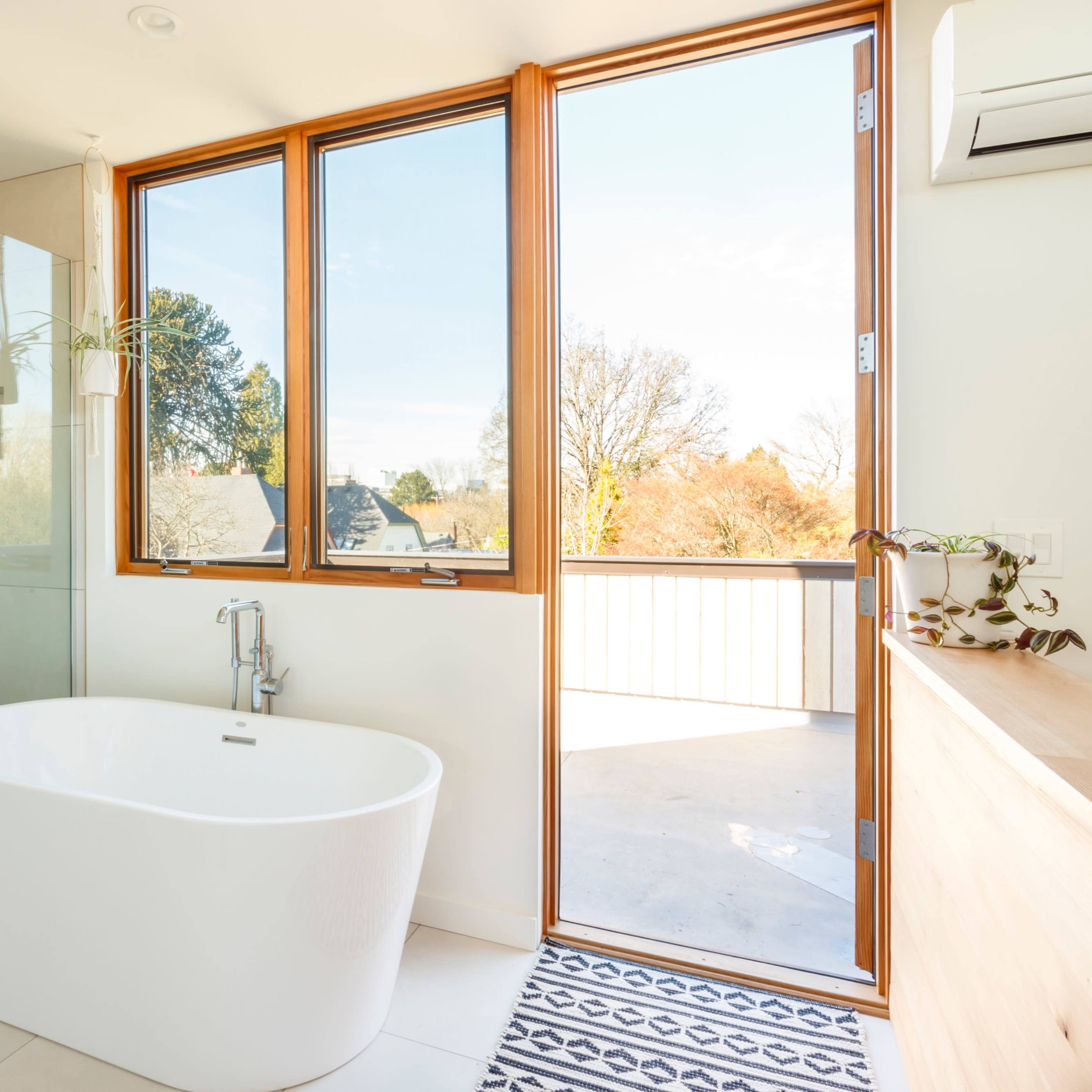A modern bathroom featuring a freestanding white bathtub, a glass shower enclosure, and large windows that provide natural light and an outdoor view.
