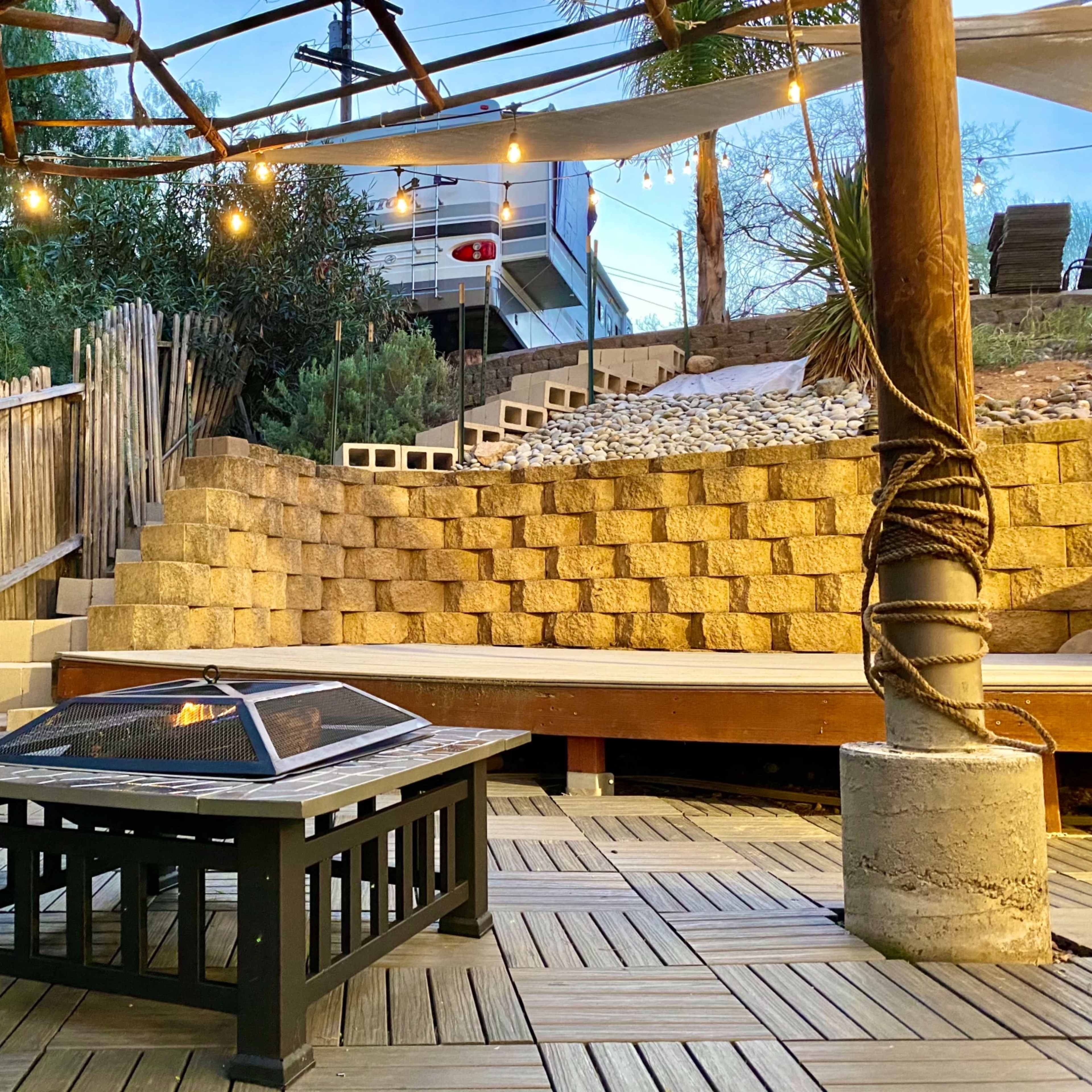 A wooden outdoor patio featuring a fire pit table, surrounded by stone steps and a wooden seating area, with string lights overhead.