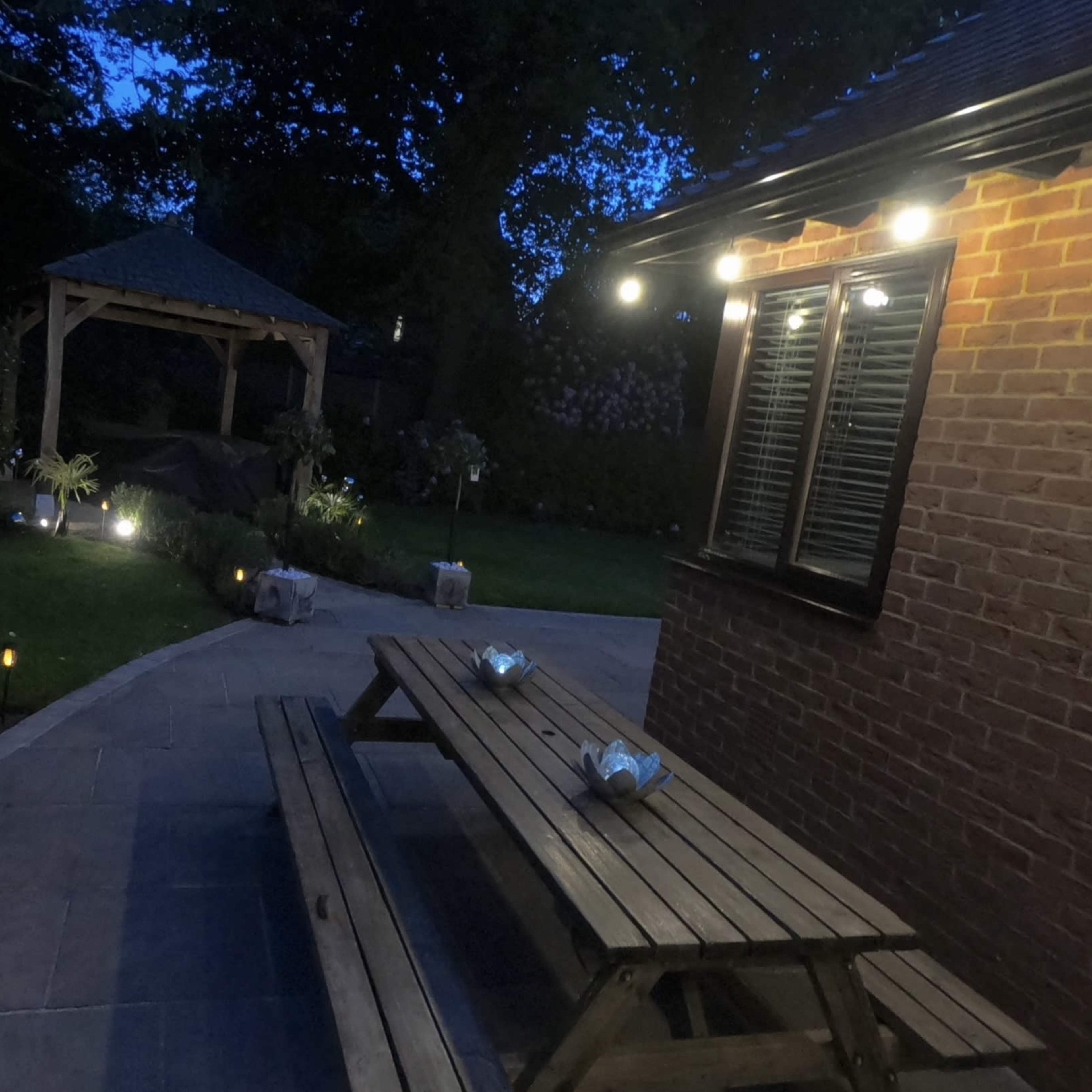 A wooden picnic table is positioned next to a well-lit brick building, with a gazebo visible in the softly illuminated garden at dusk.
