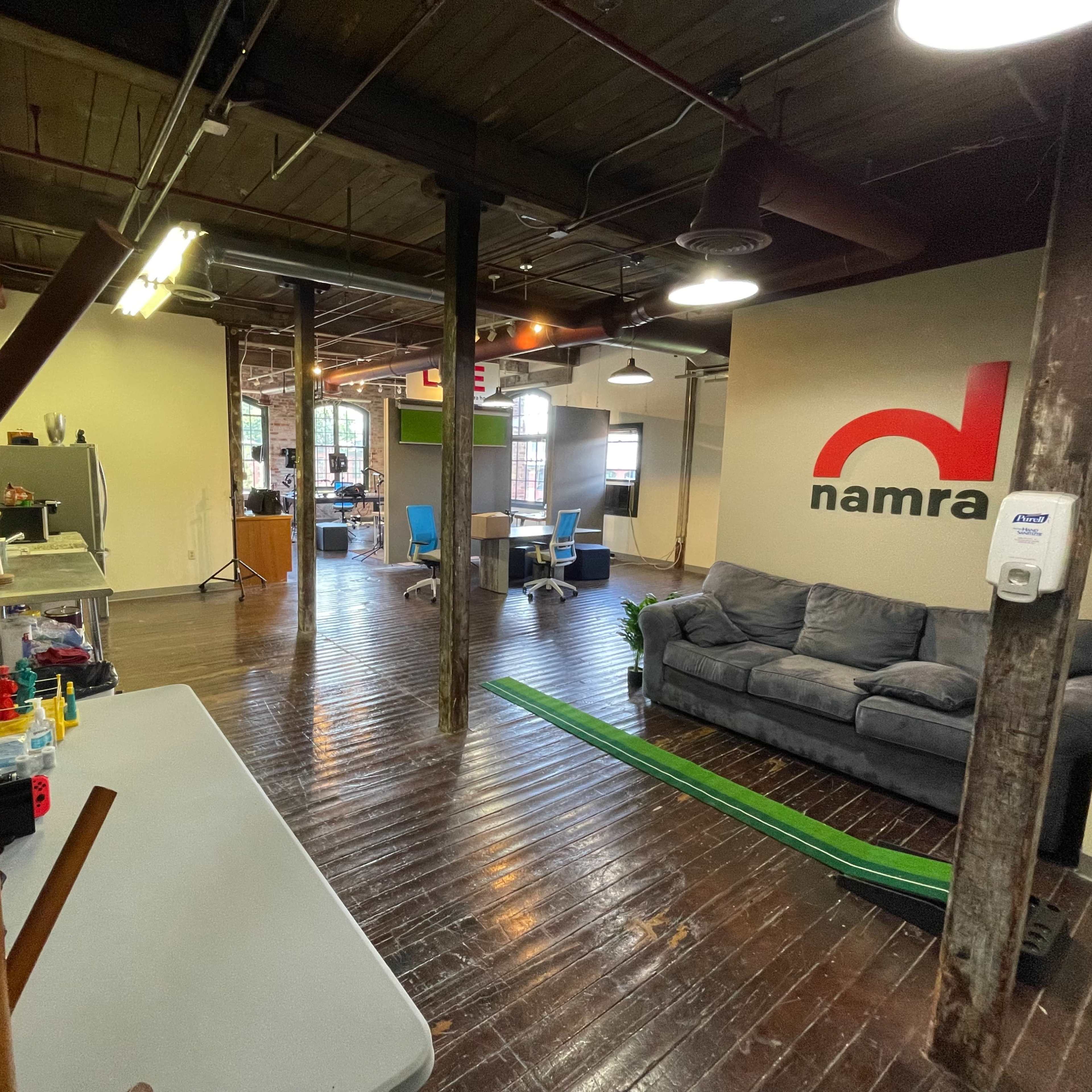 An office interior featuring a gray couch, a wooden coat rack, and a green track on the floor, with a large wall logo for "namra" in red.