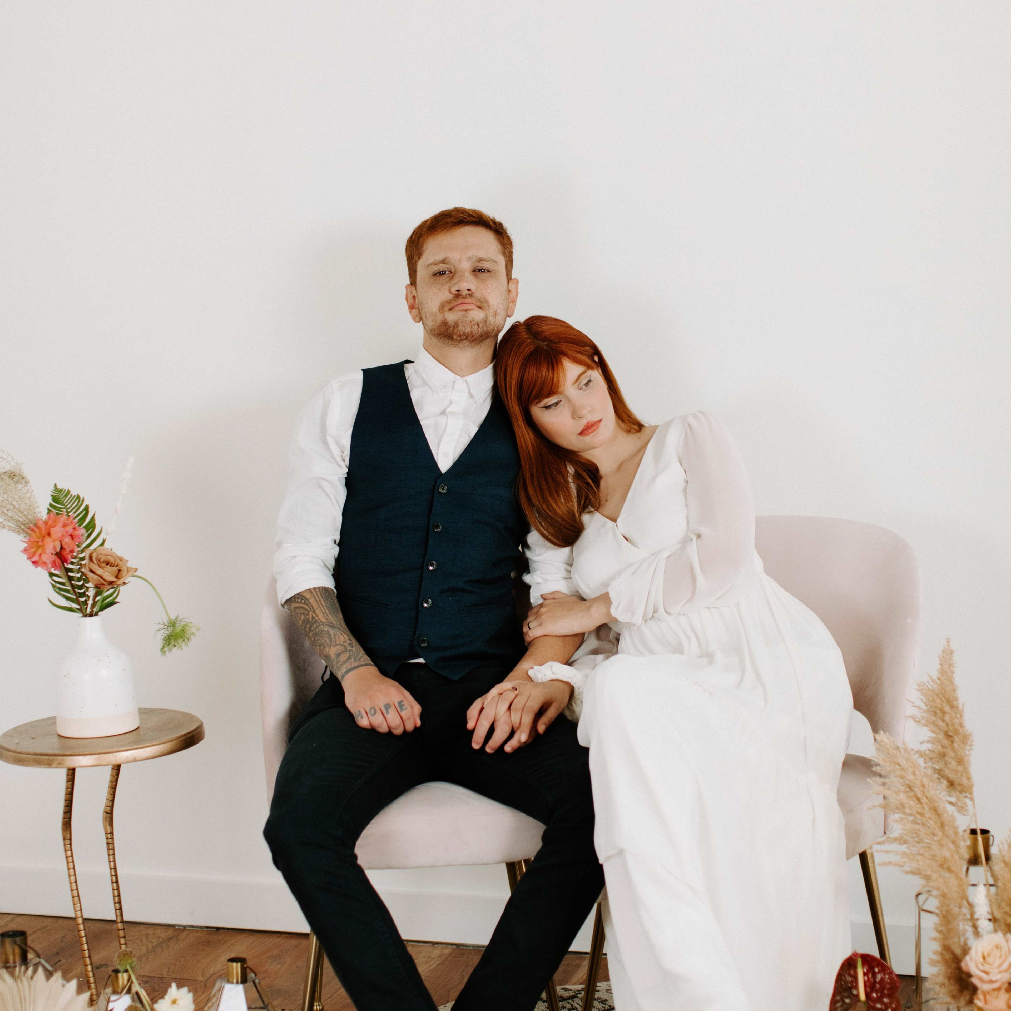 A couple sits closely together on a pink chair, with the woman resting her head on the man's shoulder in a softly decorated indoor setting.