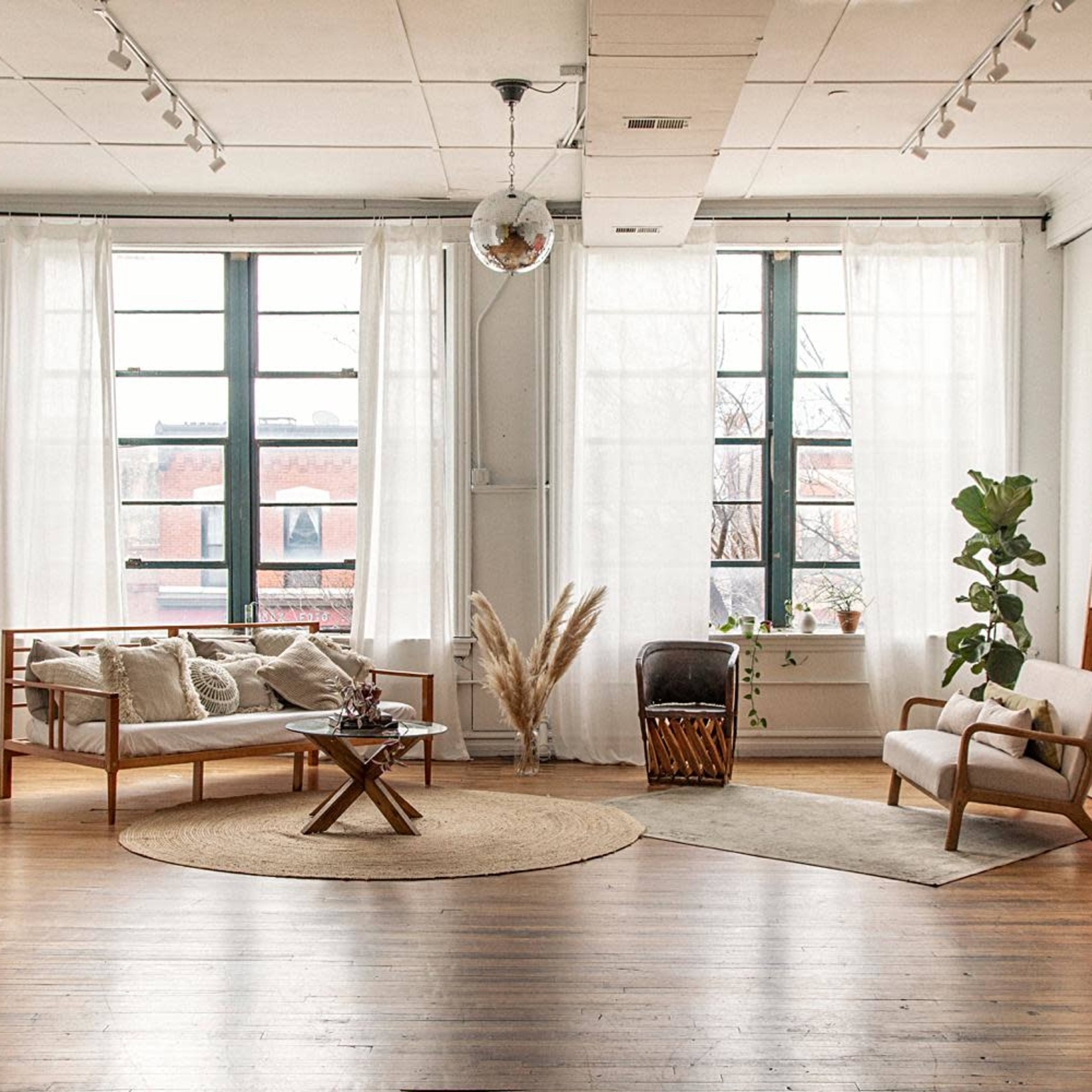 A spacious, well-lit room with large windows, featuring a light-colored sofa, a round coffee table, a chair, and a large piece of abstract art on the wall.