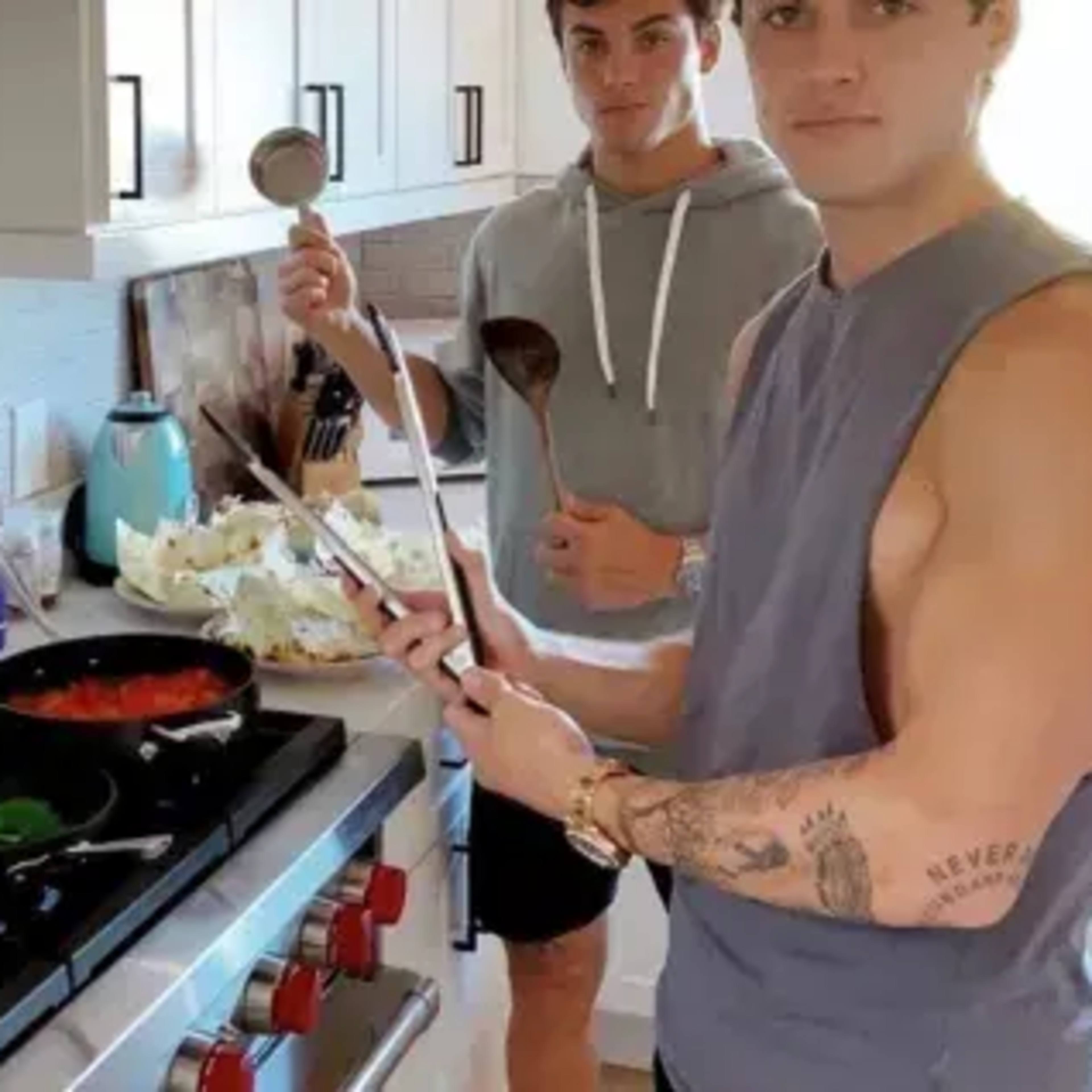 Two young men are cooking in a modern kitchen, with one stirring a pot on the stove while the other holds a wooden spoon and a ladle.