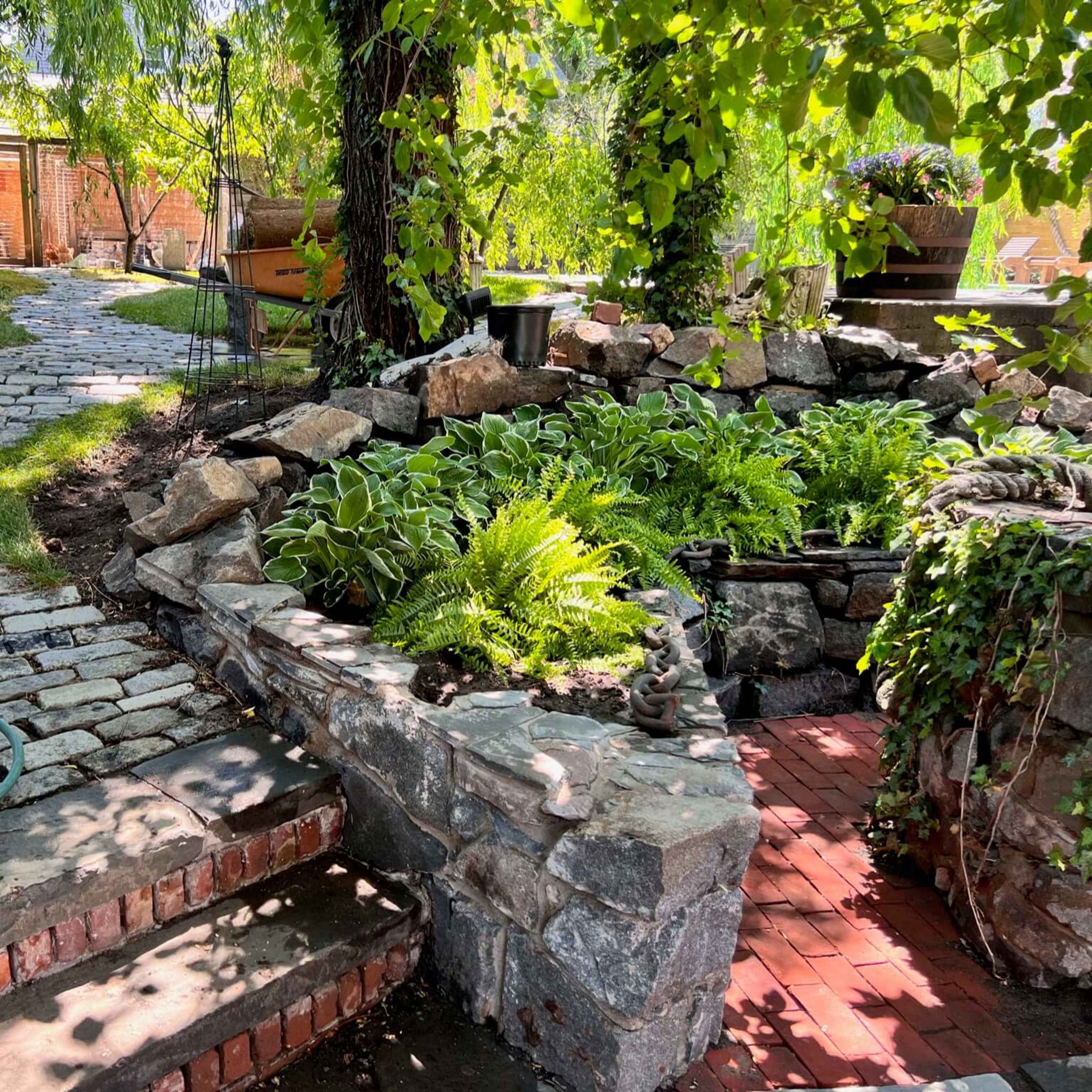 A stone pathway winds through a landscaped garden featuring a rock-bordered plant bed with ferns and hostas.