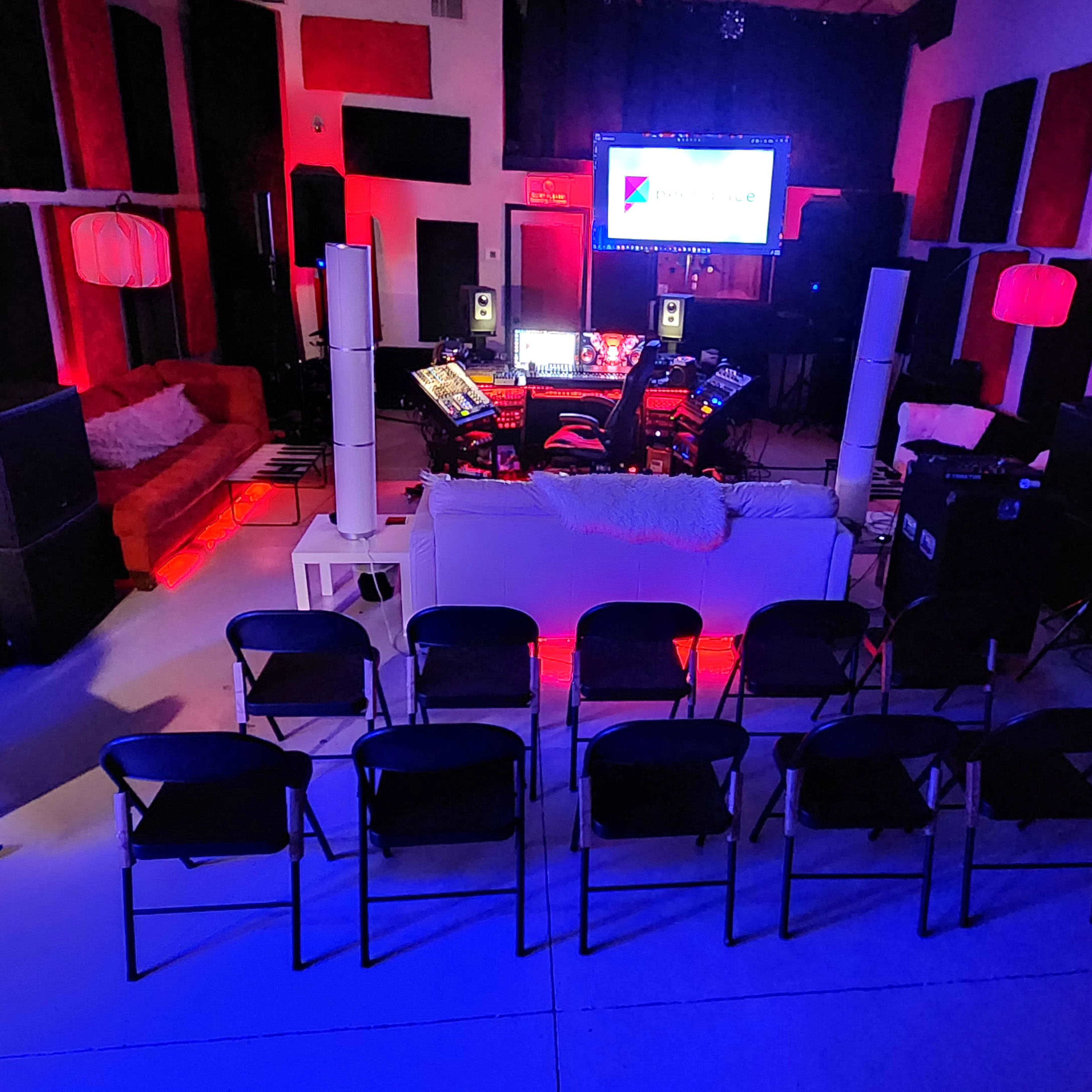 A music studio setup with seating arranged in front of a screen, surrounded by various audio equipment and colorful lighting.