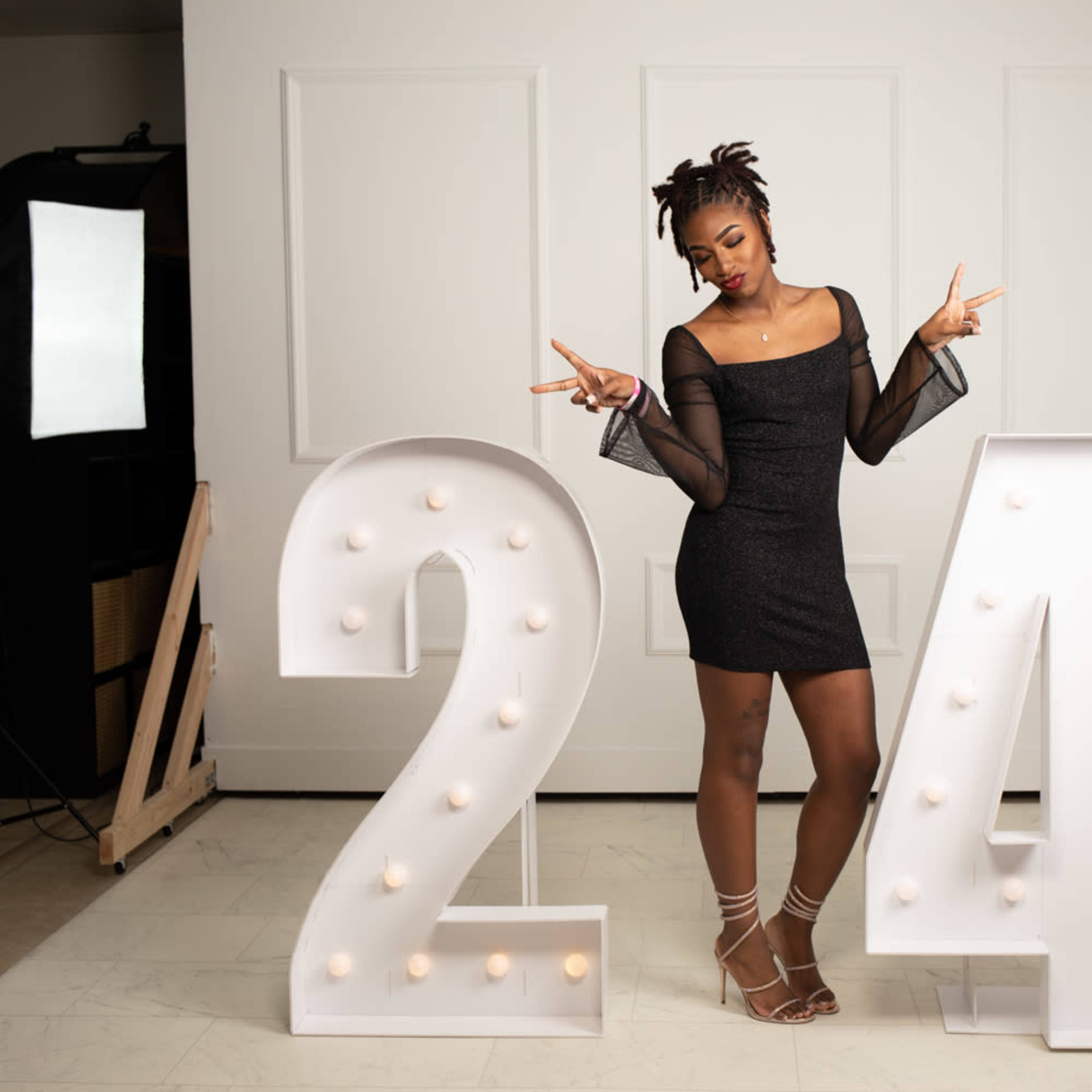 A woman poses between large illuminated numbers "24" while wearing a black dress in a well-lit studio setting.
