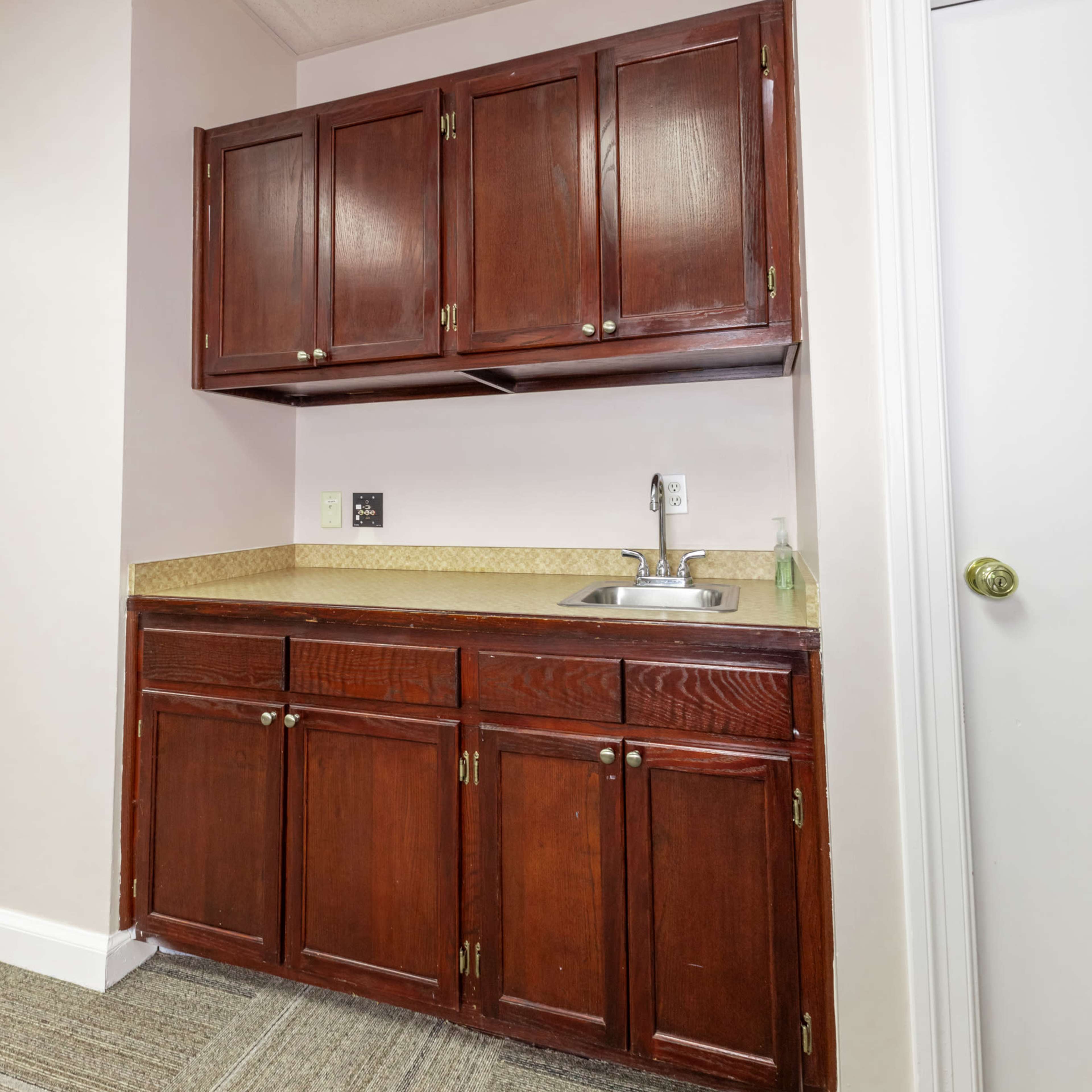 A small kitchen area featuring dark wooden cabinets above a countertop with a sink and a white door on the right.