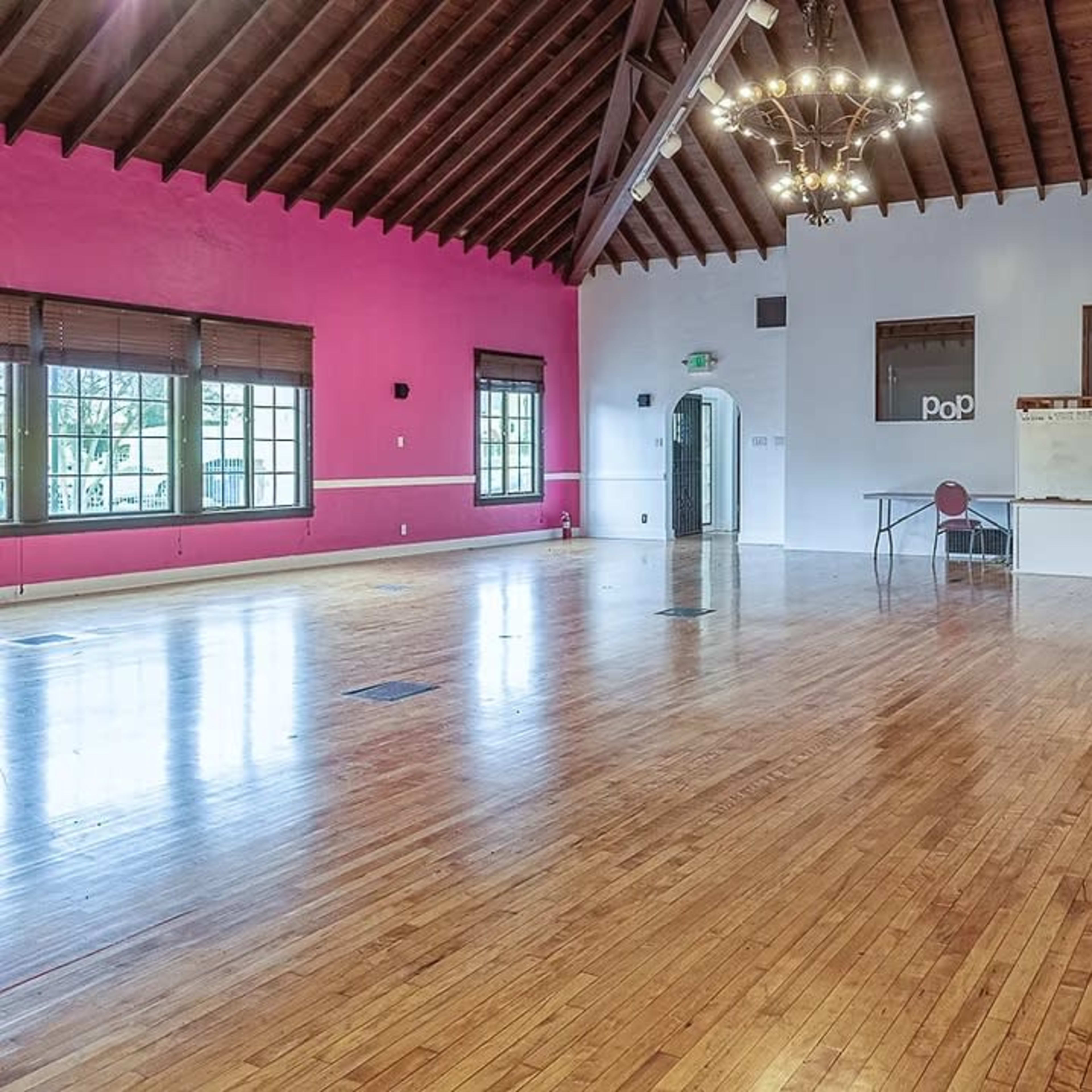 A spacious room with wooden floors, high ceilings, and a mix of pink and white walls, along with a table and chairs set up in one corner.