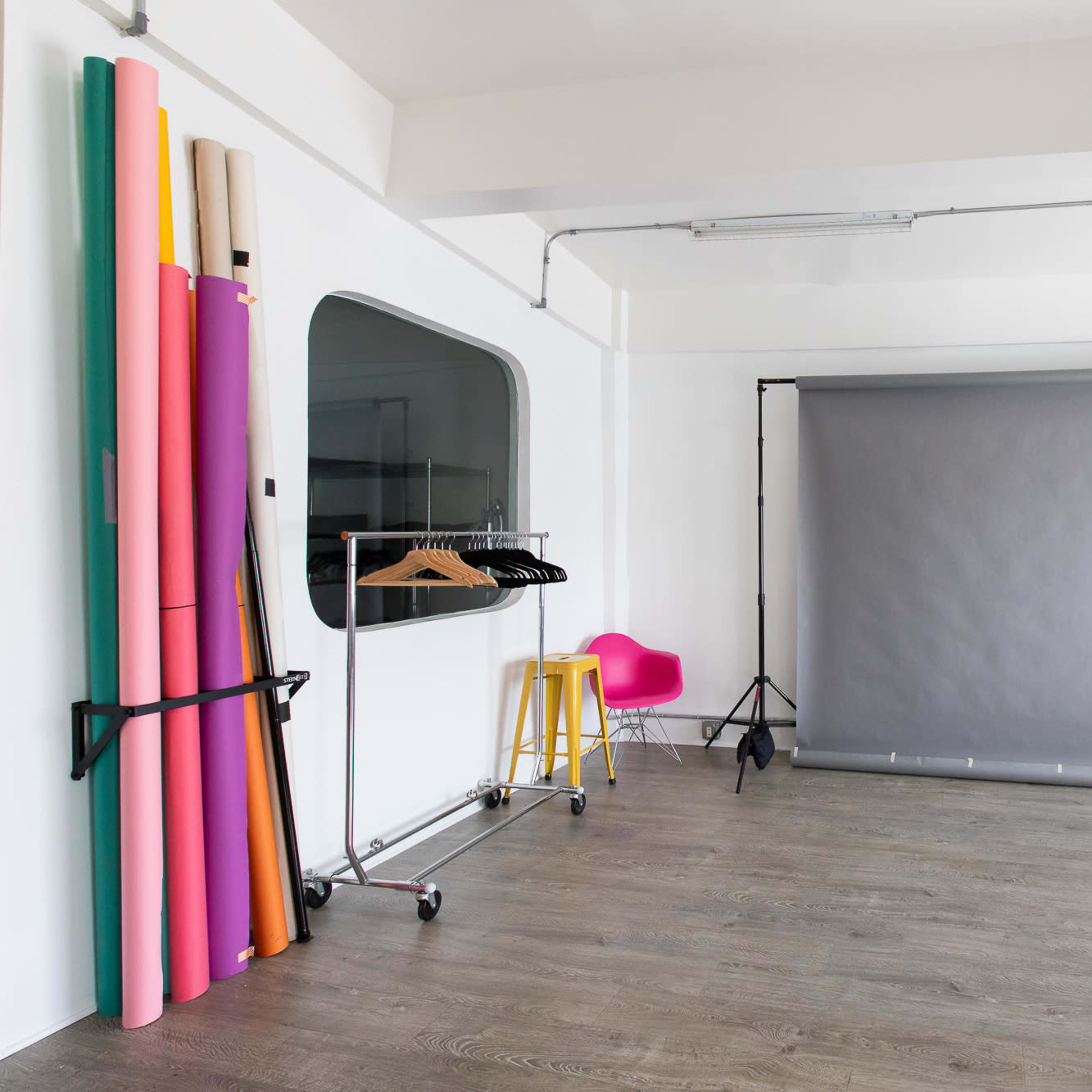 A photography studio featuring a gray backdrop, colorful rolls of paper, clothing on a rack, and potted plants.