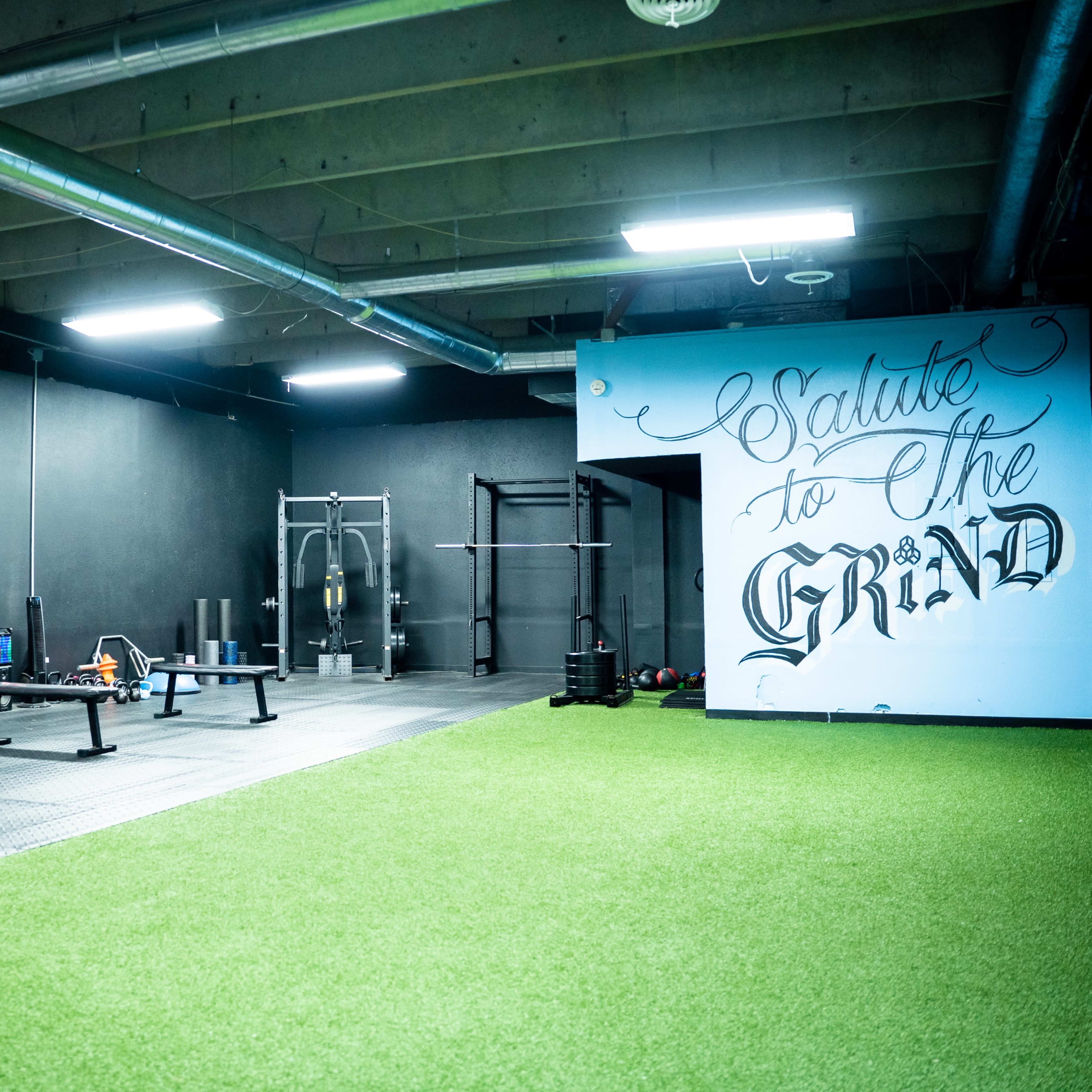 A fitness studio features various weightlifting equipment and a turf area, with one wall adorned with a mural that reads "Salute to the Grind."