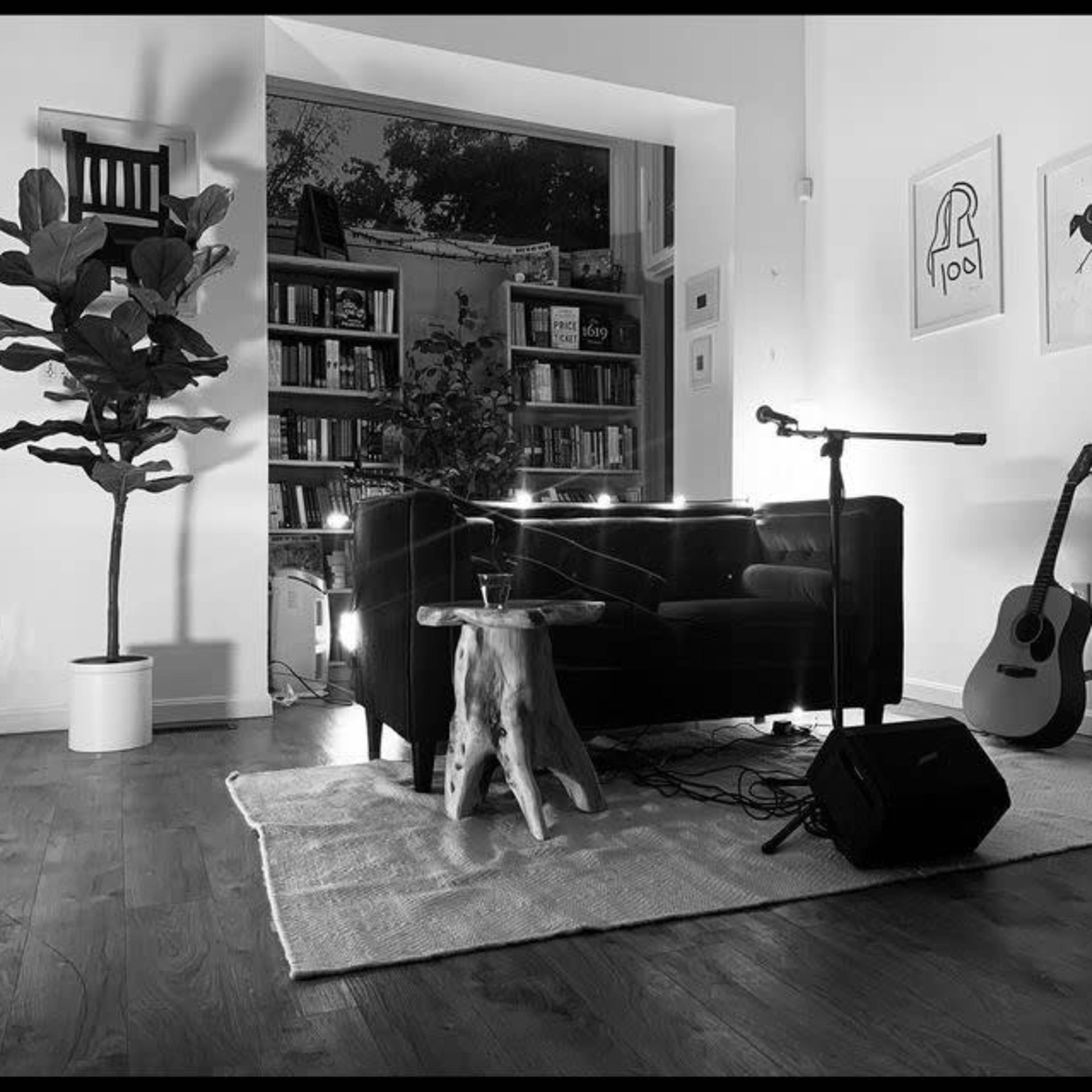 A minimalist living room with a black couch, a small round table made of wood, a microphone on a stand, a guitar leaning against the wall, and bookshelves in the background.