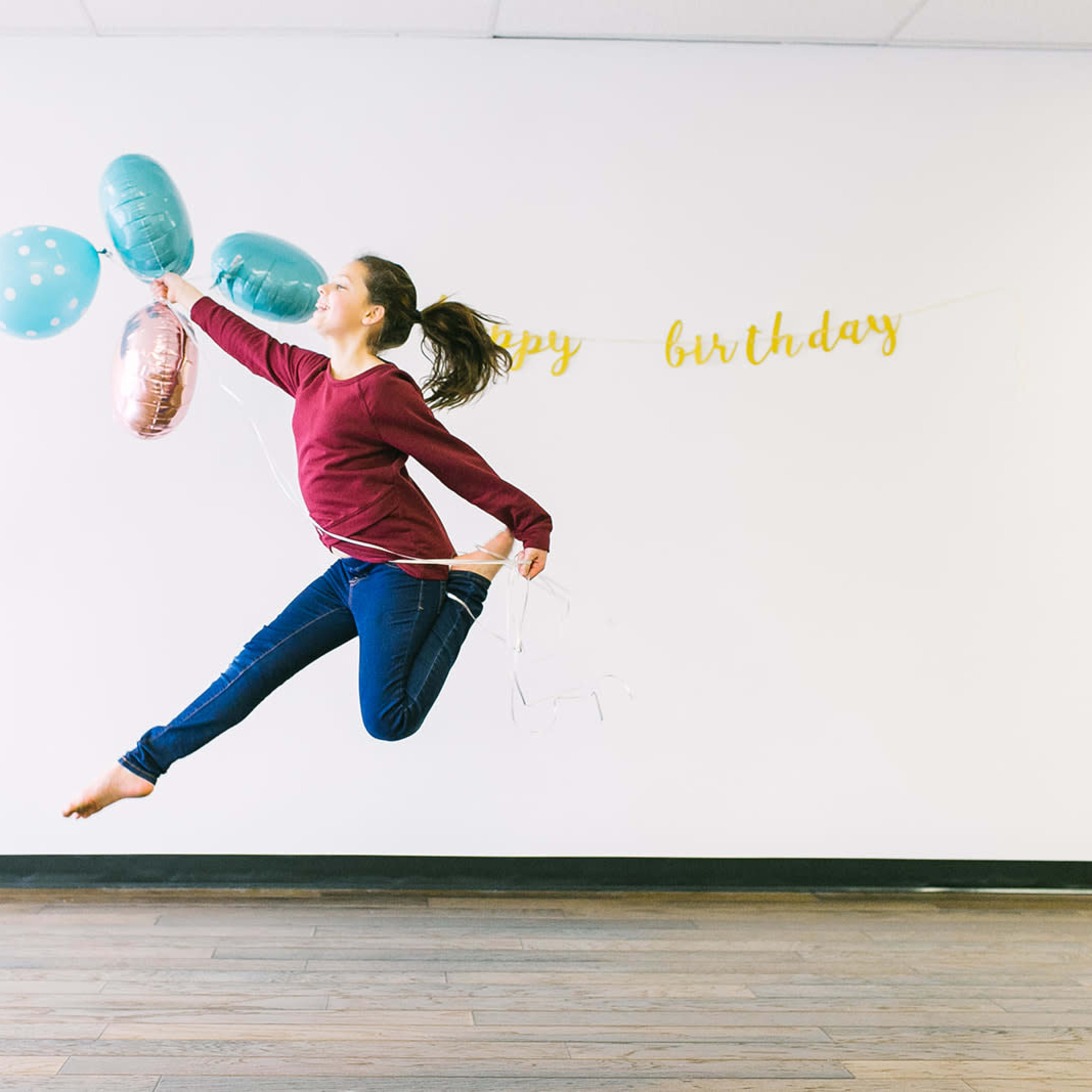 A young girl jumps joyfully while holding a bunch of balloons against a wall decorated with a "Happy Birthday" banner.
