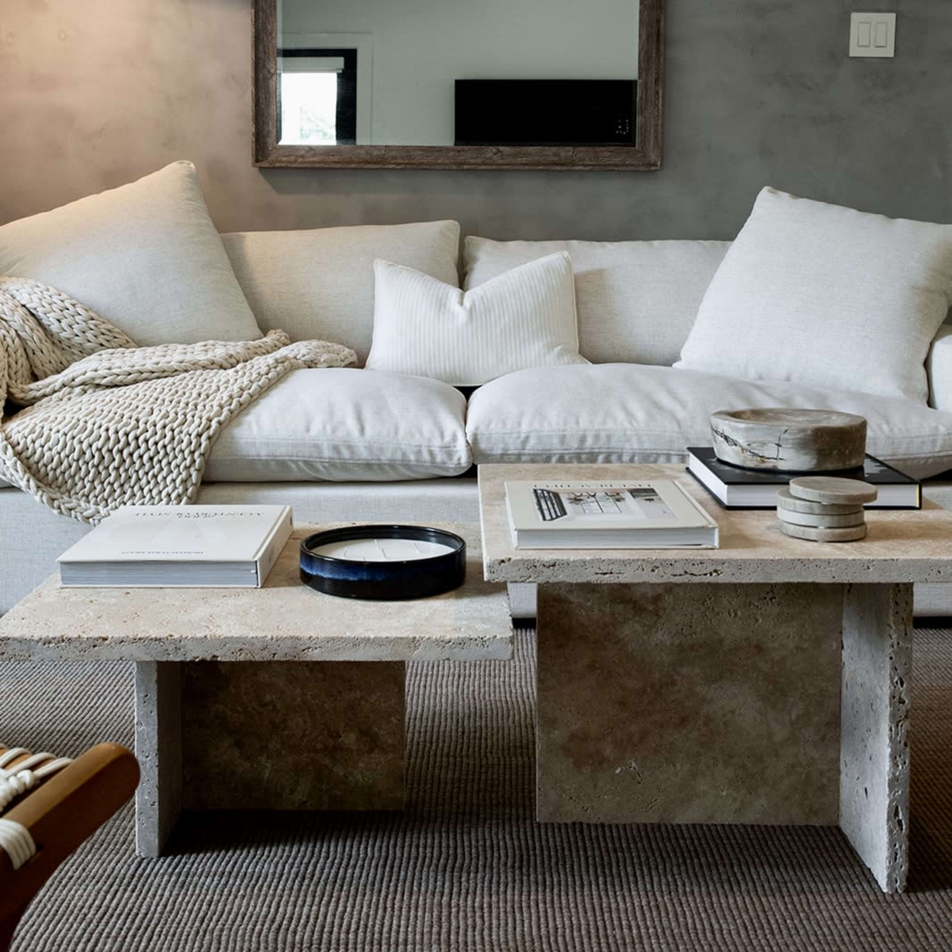 A light-colored sofa with assorted cushions sits in front of a textured gray wall, accompanied by a minimalist coffee table displaying books and decorative objects.