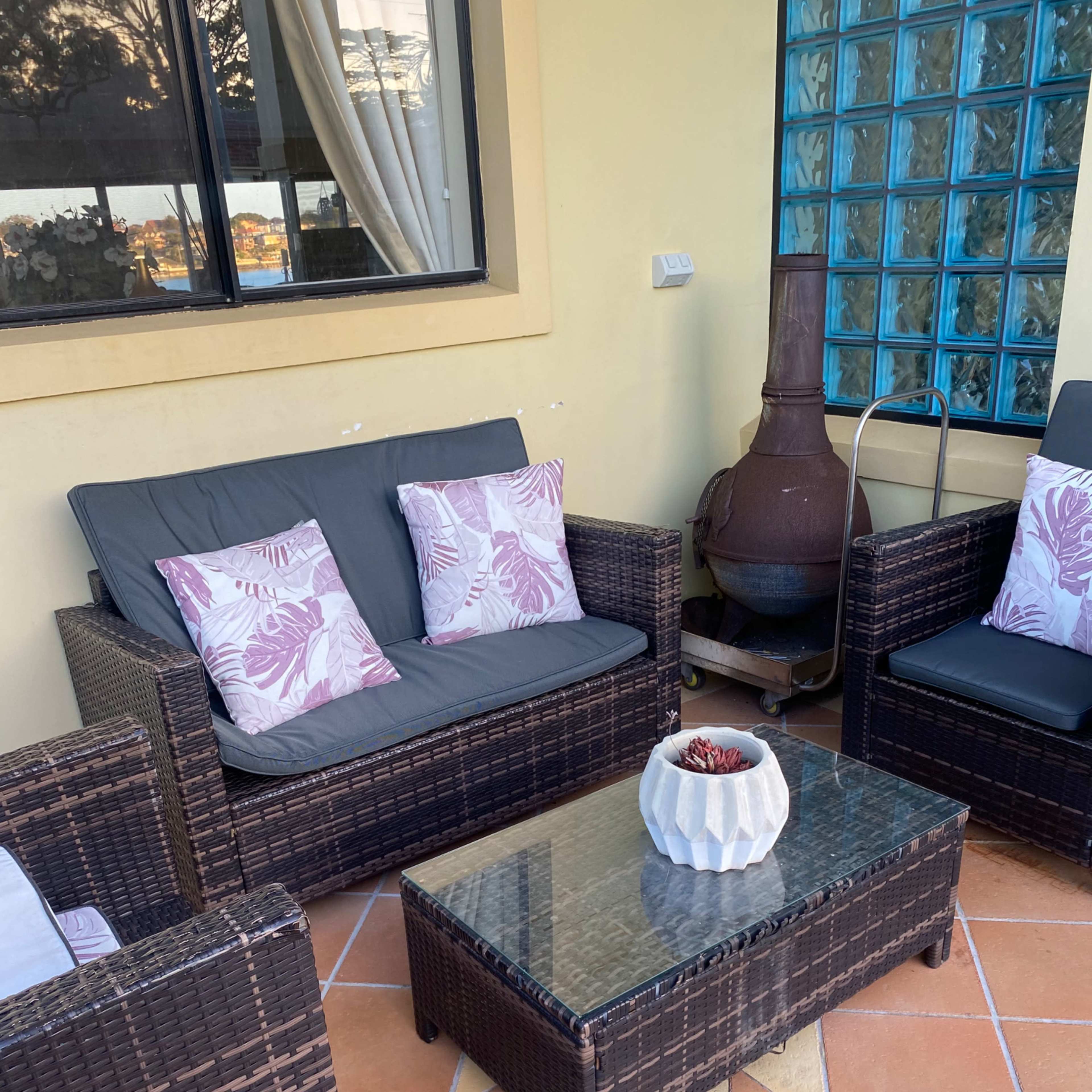 An outdoor seating area featuring a rattan sofa set with cushions, a glass coffee table, and a chiminea beside a wall with a large window.