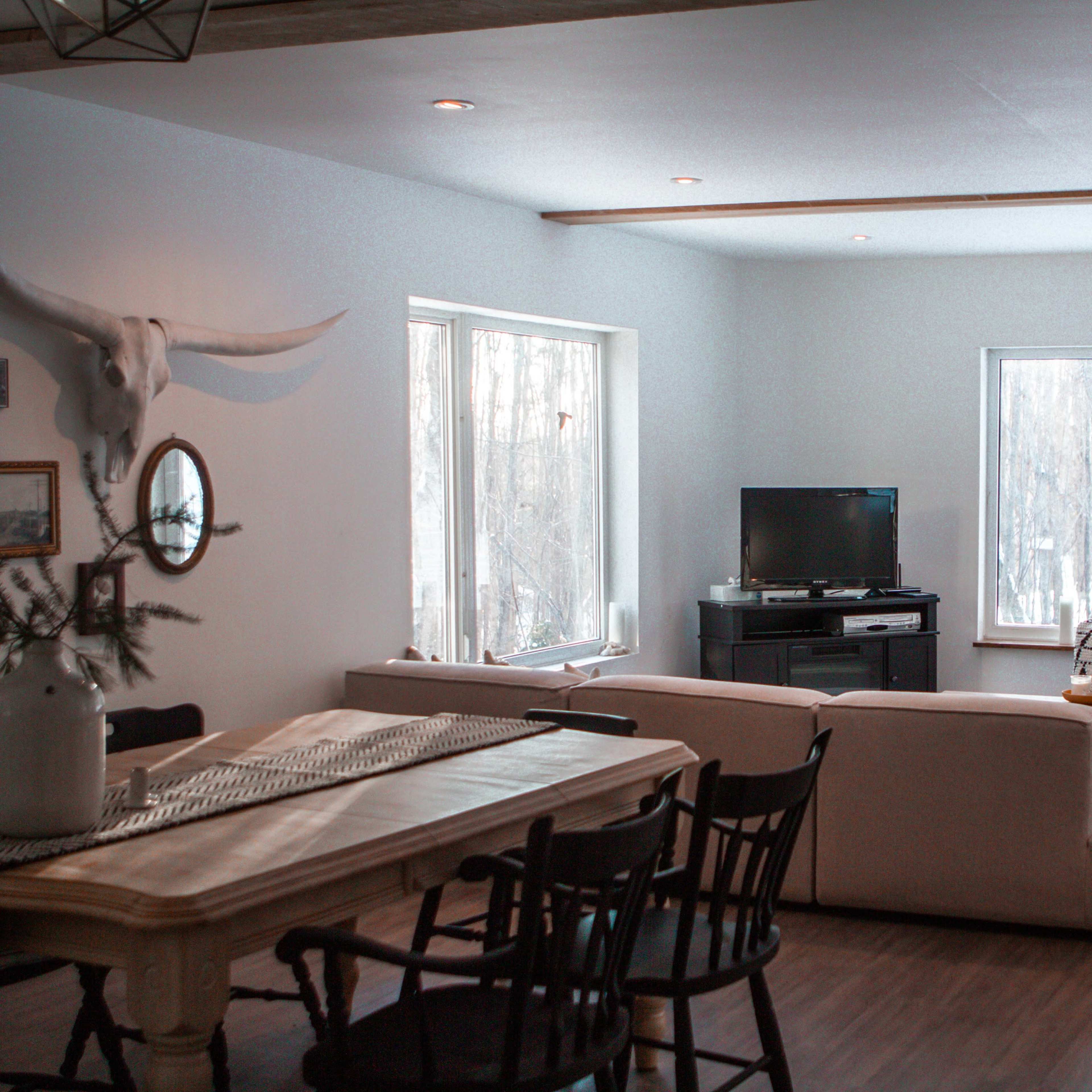 A modern living room with a white couch, a wooden dining table, and large windows overlooking a snowy landscape.