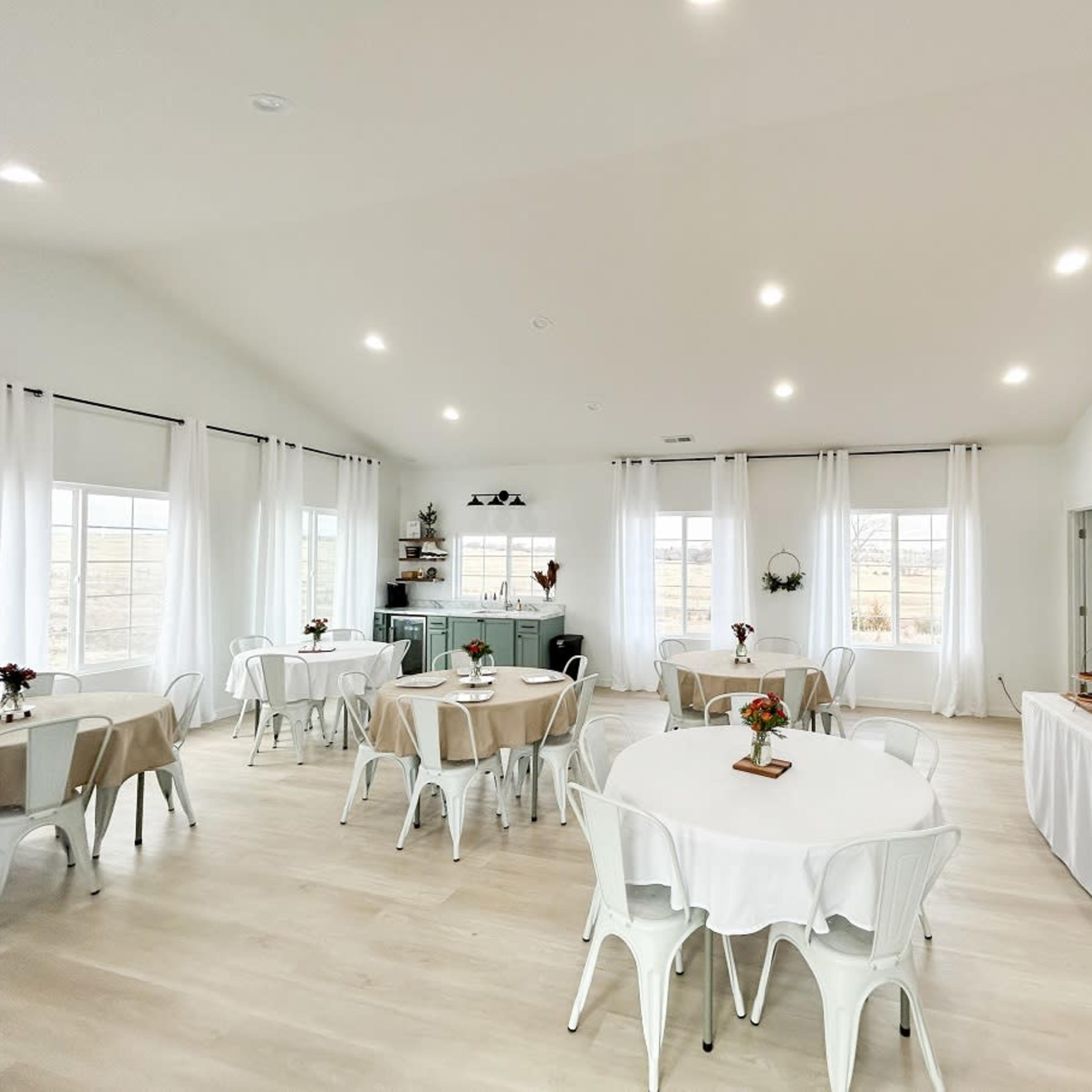A bright, airy event space with several round tables set for guests, large windows, and a decorated service area featuring a coffee station.