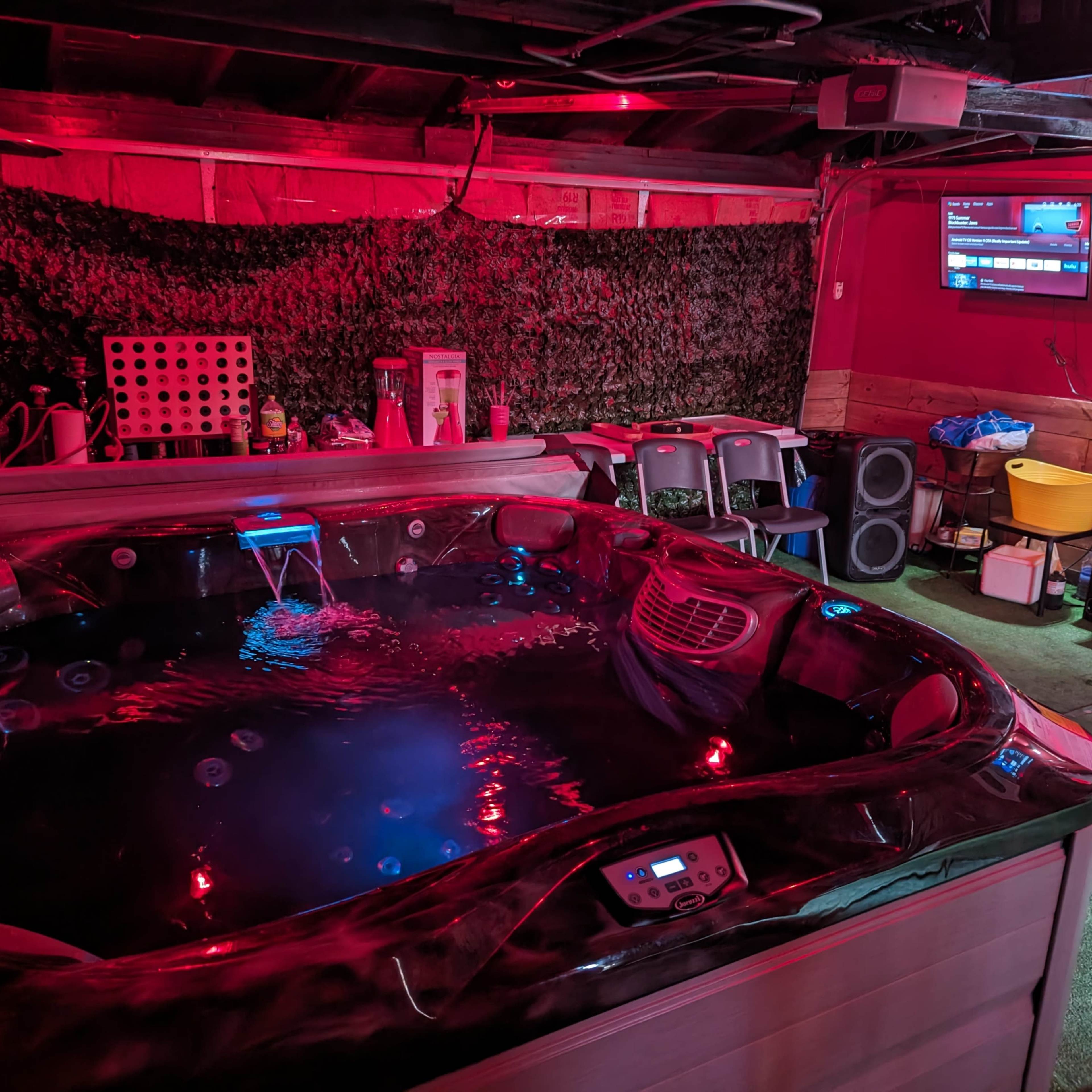 A hot tub in a dimly lit basement with red lighting and a television mounted on the wall.
